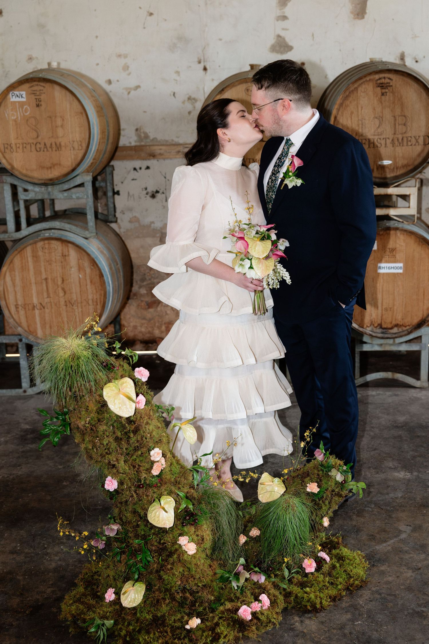 Bride and groom kissing at winery wedding venue surrounded by wine barrels and floral moss arrangement.