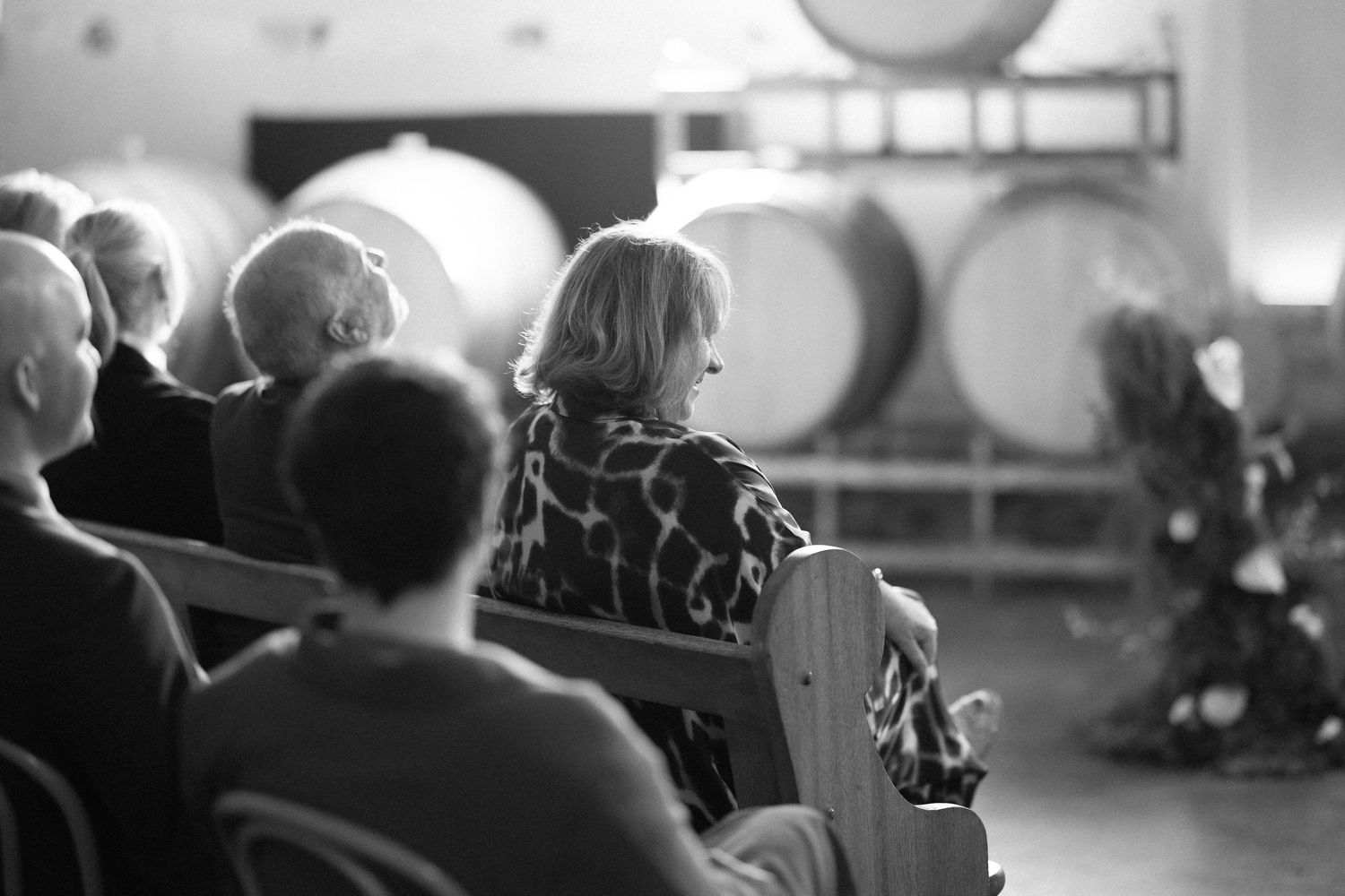Black and white photo of wedding guests seated on wooden pews in a winery ceremony space.