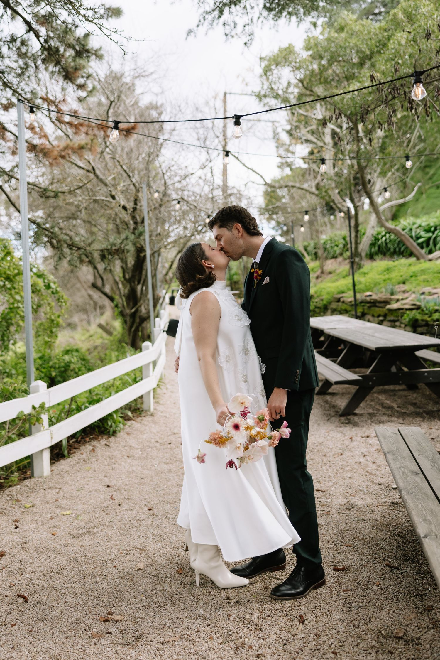 A bride and groom share a kiss outdoors at a garden wedding venue, surrounded by trees and string lights.