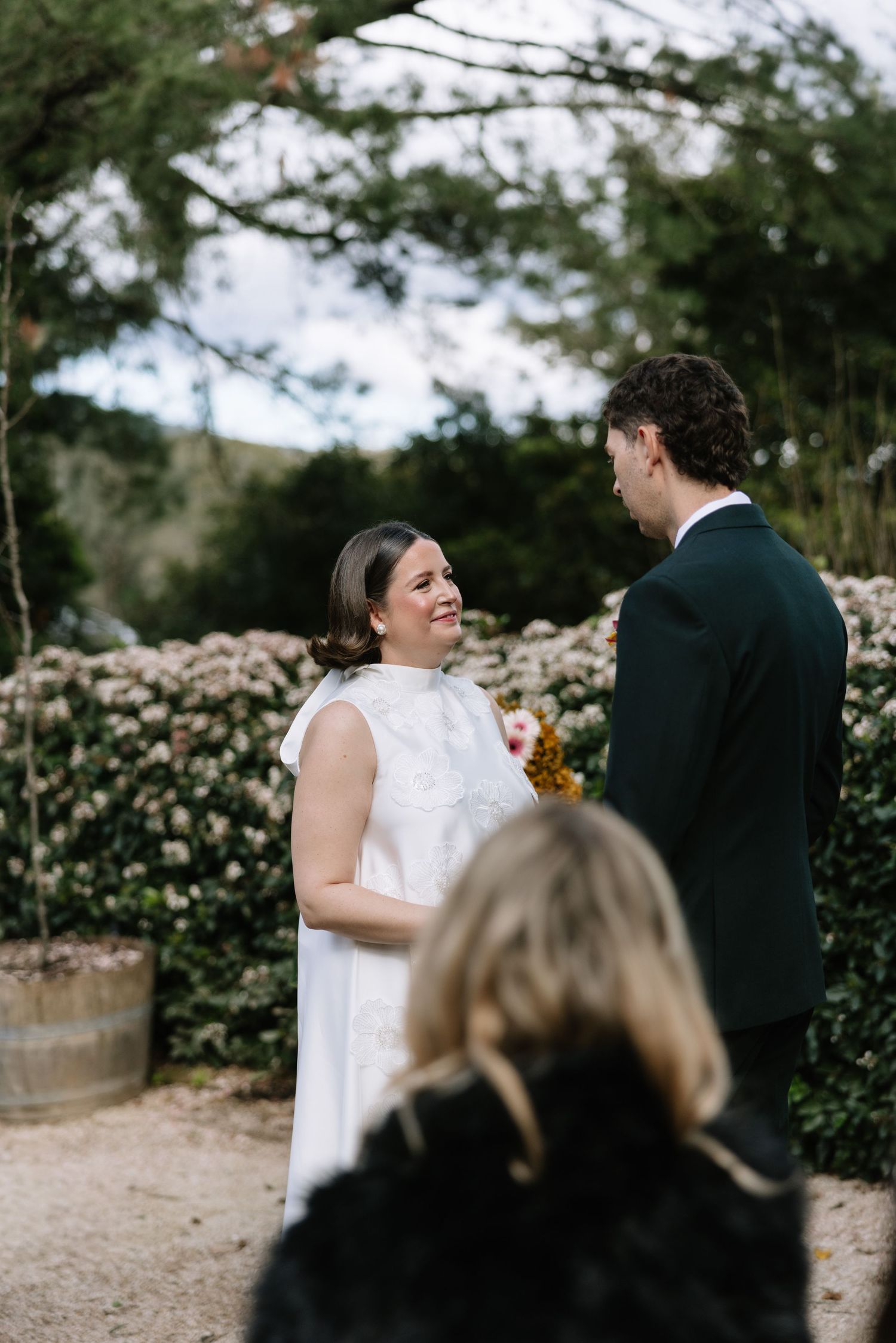 Bride smiling at groom during outdoor wedding ceremony, surrounded by lush greenery and flowering hedges.