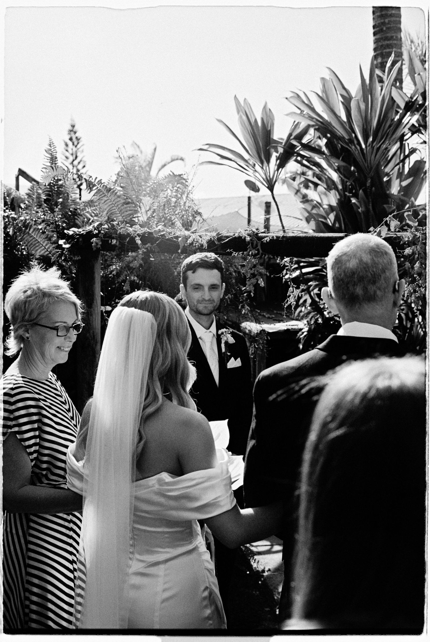 Black and white photo of a bride in veil facing groom at outdoor wedding ceremony surrounded by tropical greenery.