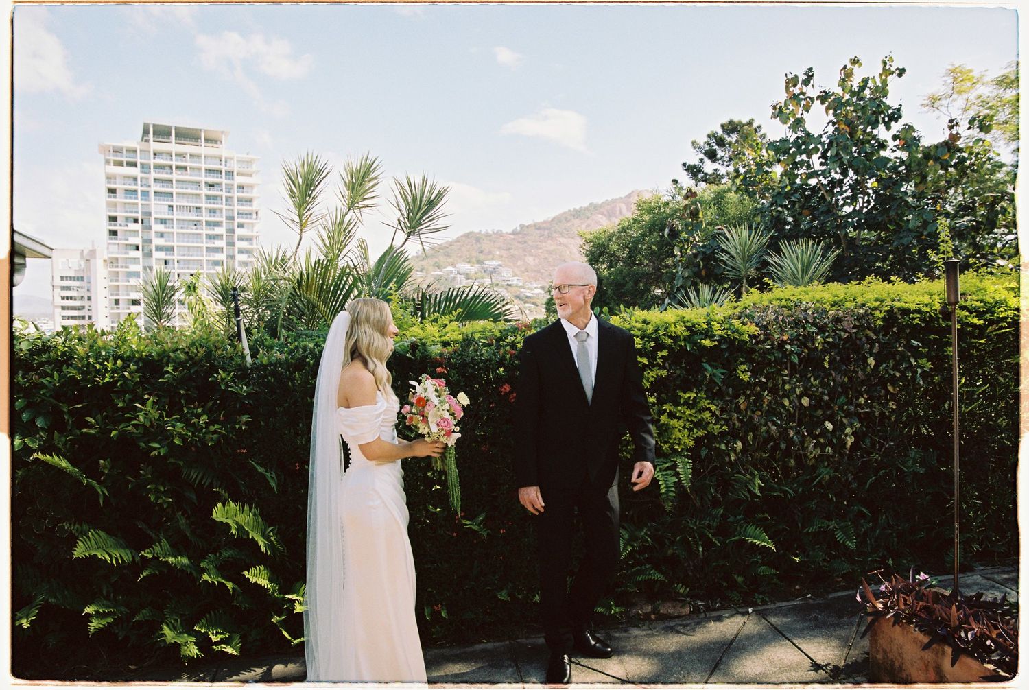 Bride in white gown with veil walks with father through lush garden with city buildings behind.