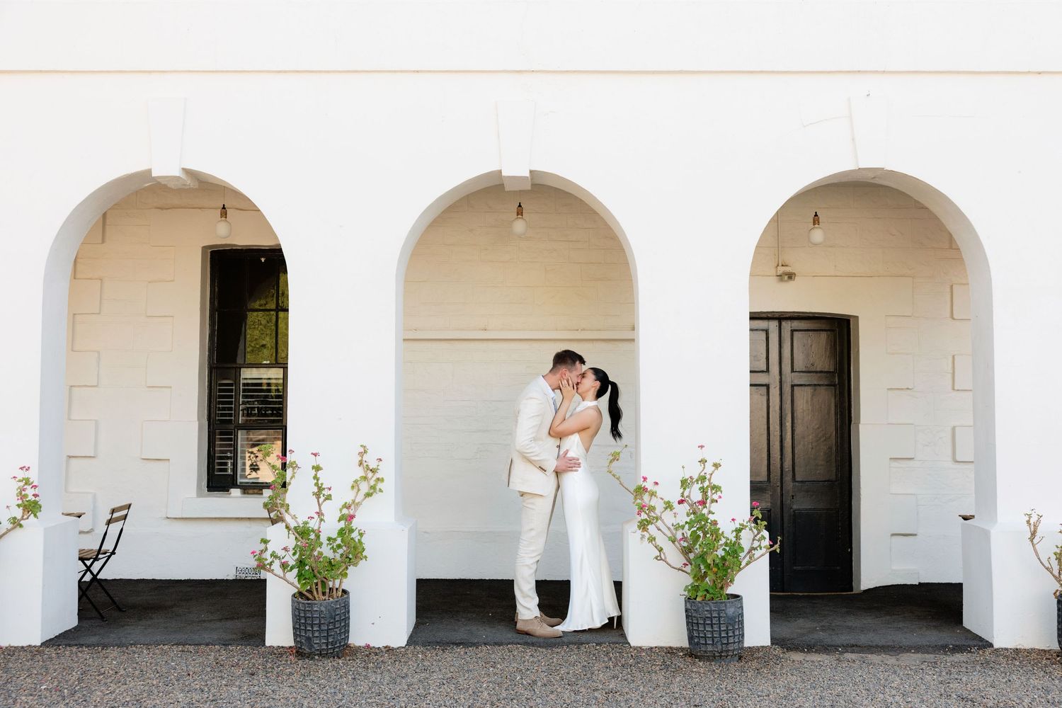 Couple sharing a romantic kiss under white arched colonnade at elegant wedding venue.