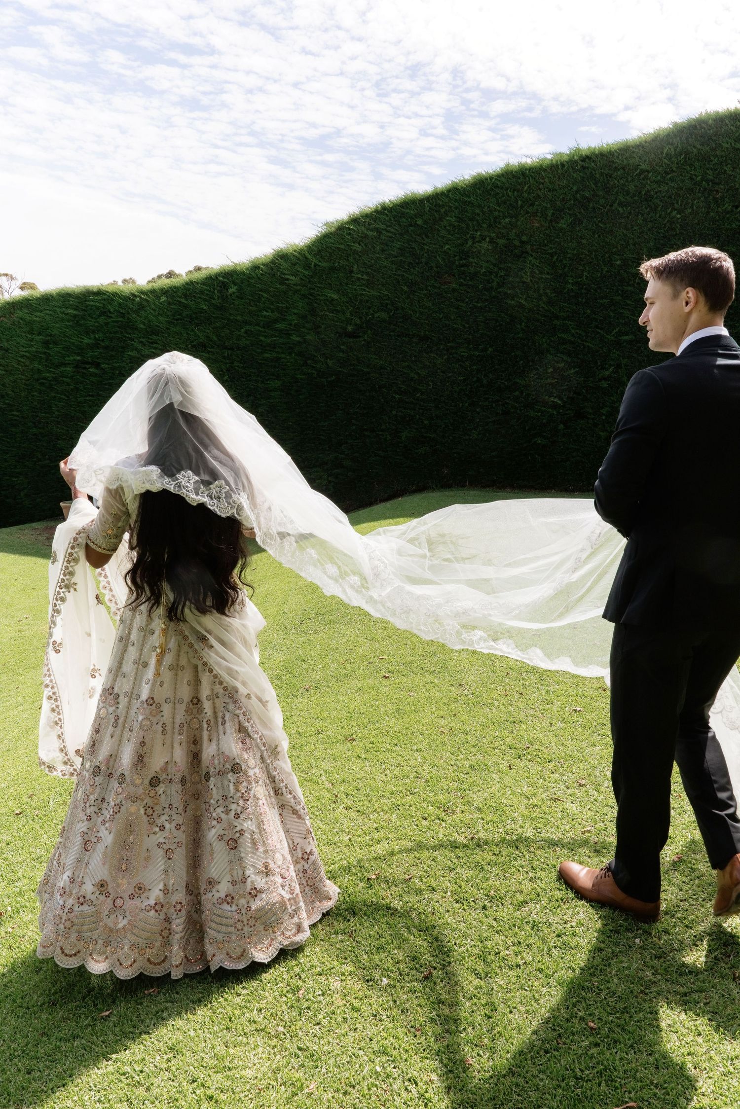 Bride in floral lehenga with flowing veil caught by wind, groom in black tuxedo, lush green hedge backdrop.