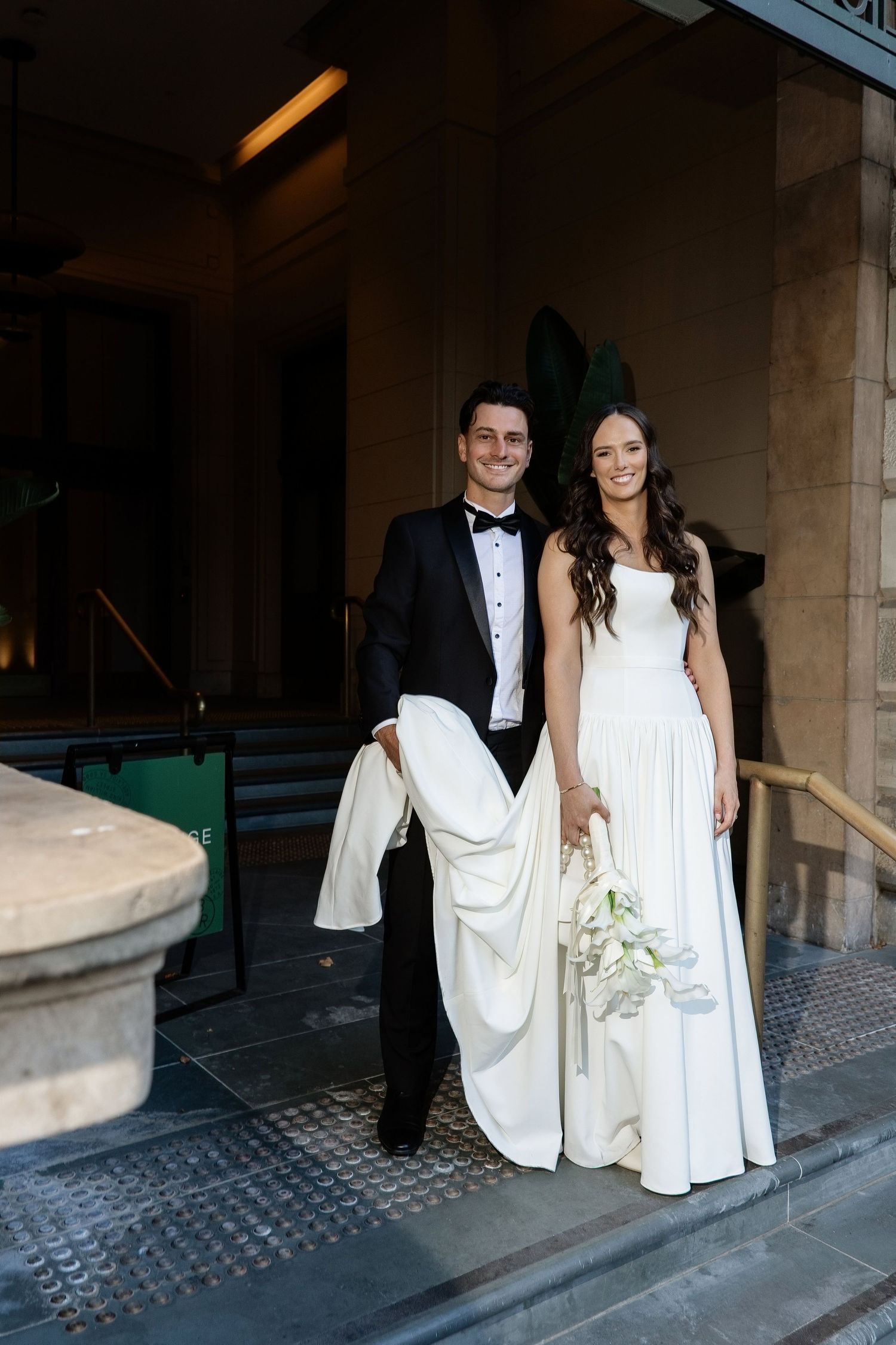 Happy bride and groom standing at venue entrance, groom carrying bride's train, bride holding white floral bouquet.