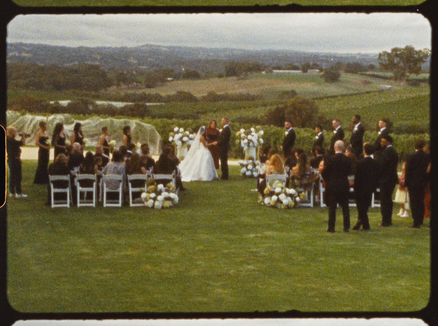 Wide aerial view of outdoor vineyard wedding ceremony with guests seated on white chairs.