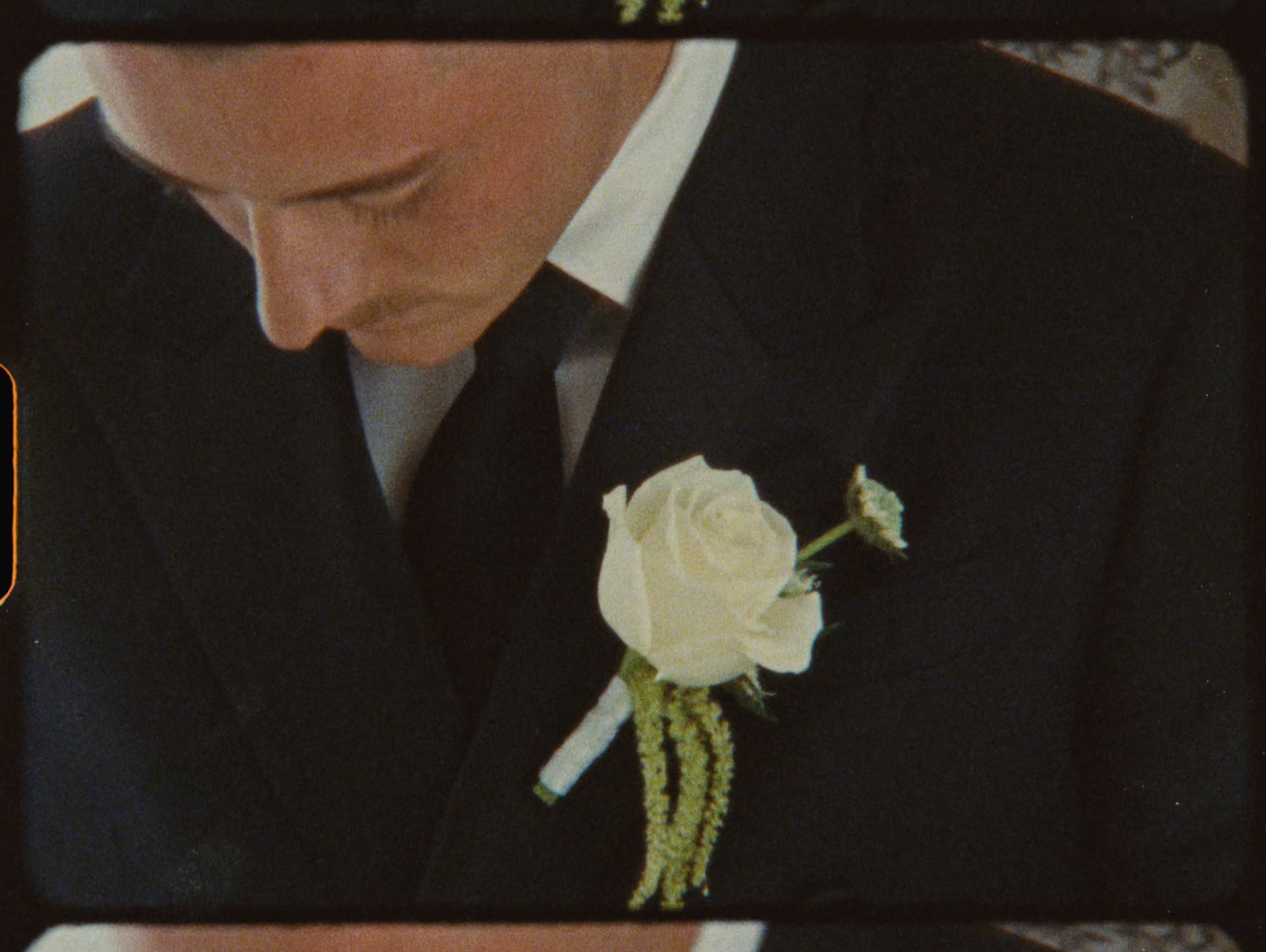 Groom in black suit with white rose boutonniere looking down at wedding ceremony.
