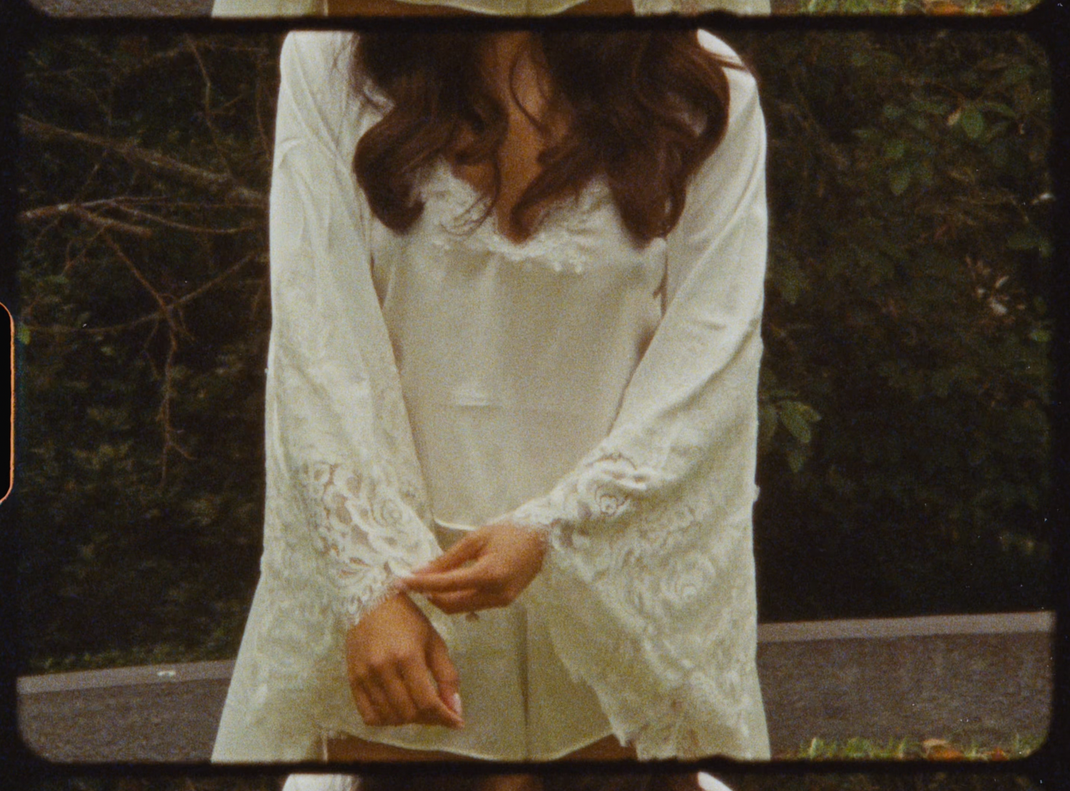 Bride in a white lace long-sleeve wedding robe holding hands in front of greenery.