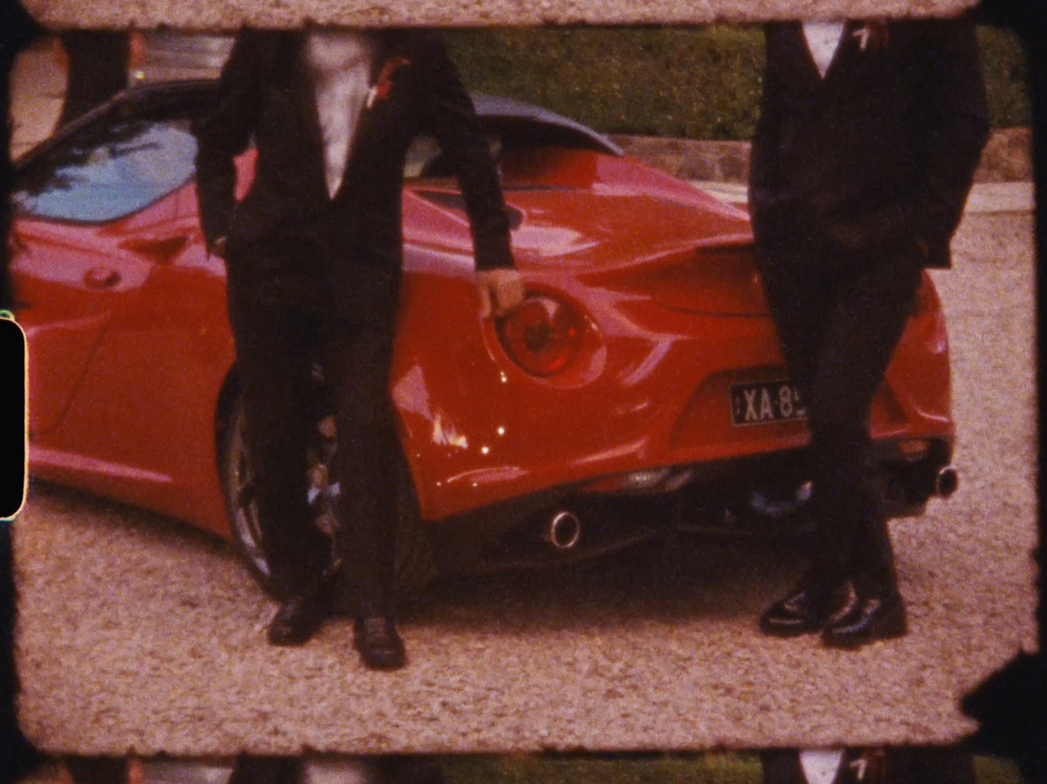 Two men in black suits standing beside a red Ferrari sports car at outdoor event.
