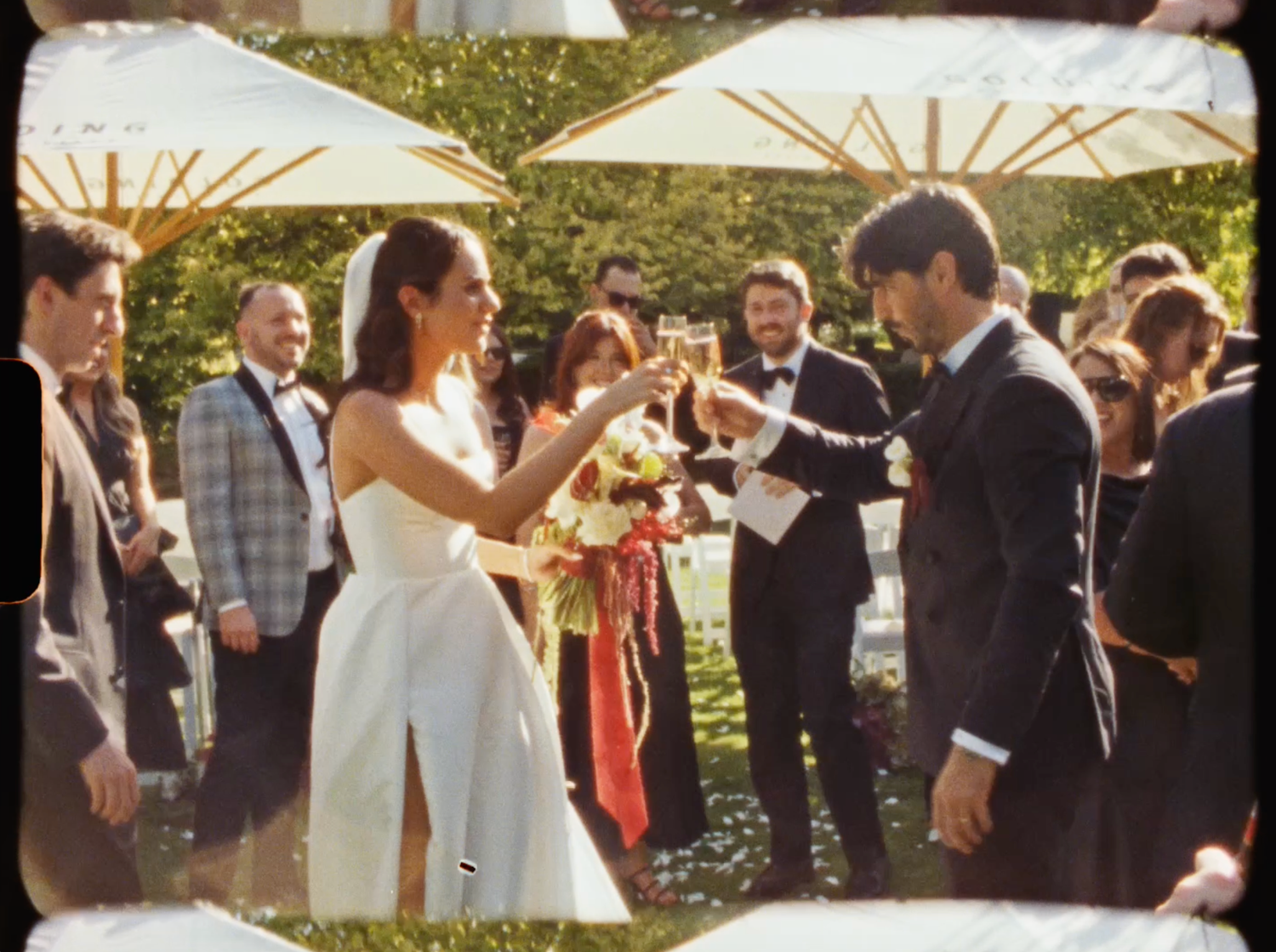 Newlyweds toast with champagne glasses surrounded by smiling guests at their outdoor wedding reception.