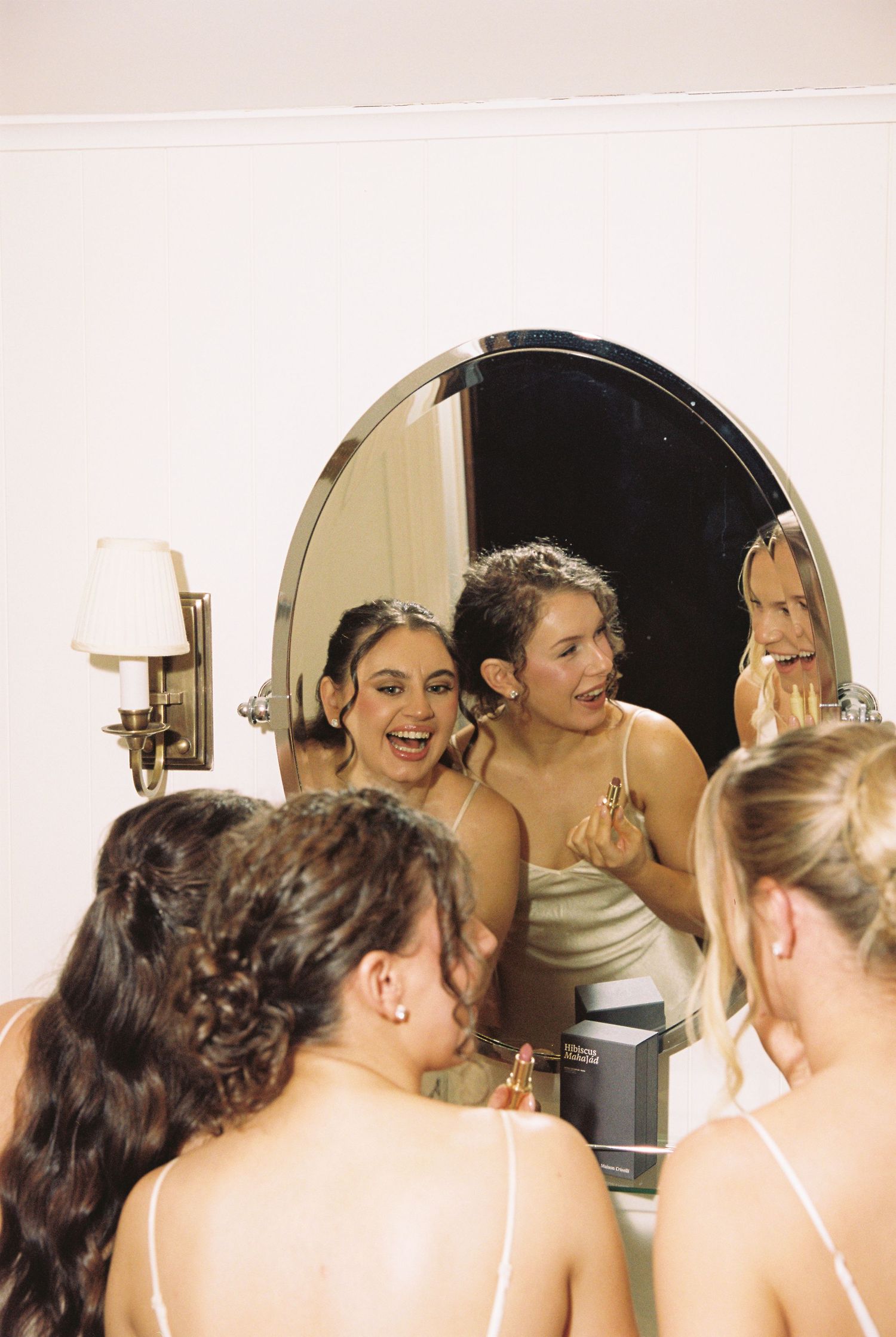 Bridesmaids in satin dresses laughing and applying makeup together in a round mirror.