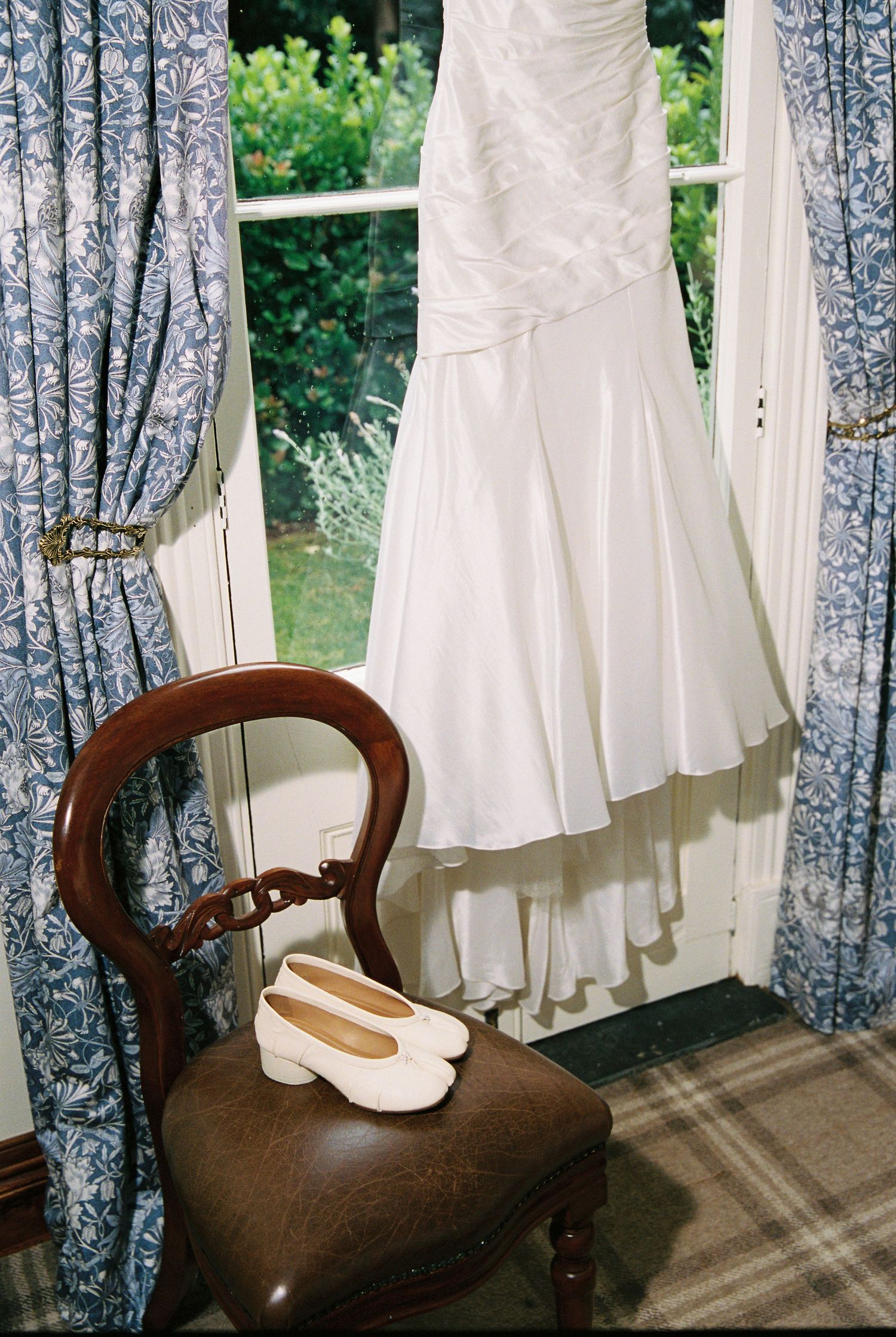 White wedding dress hanging by window with white block heel bridal shoes on vintage wooden chair.