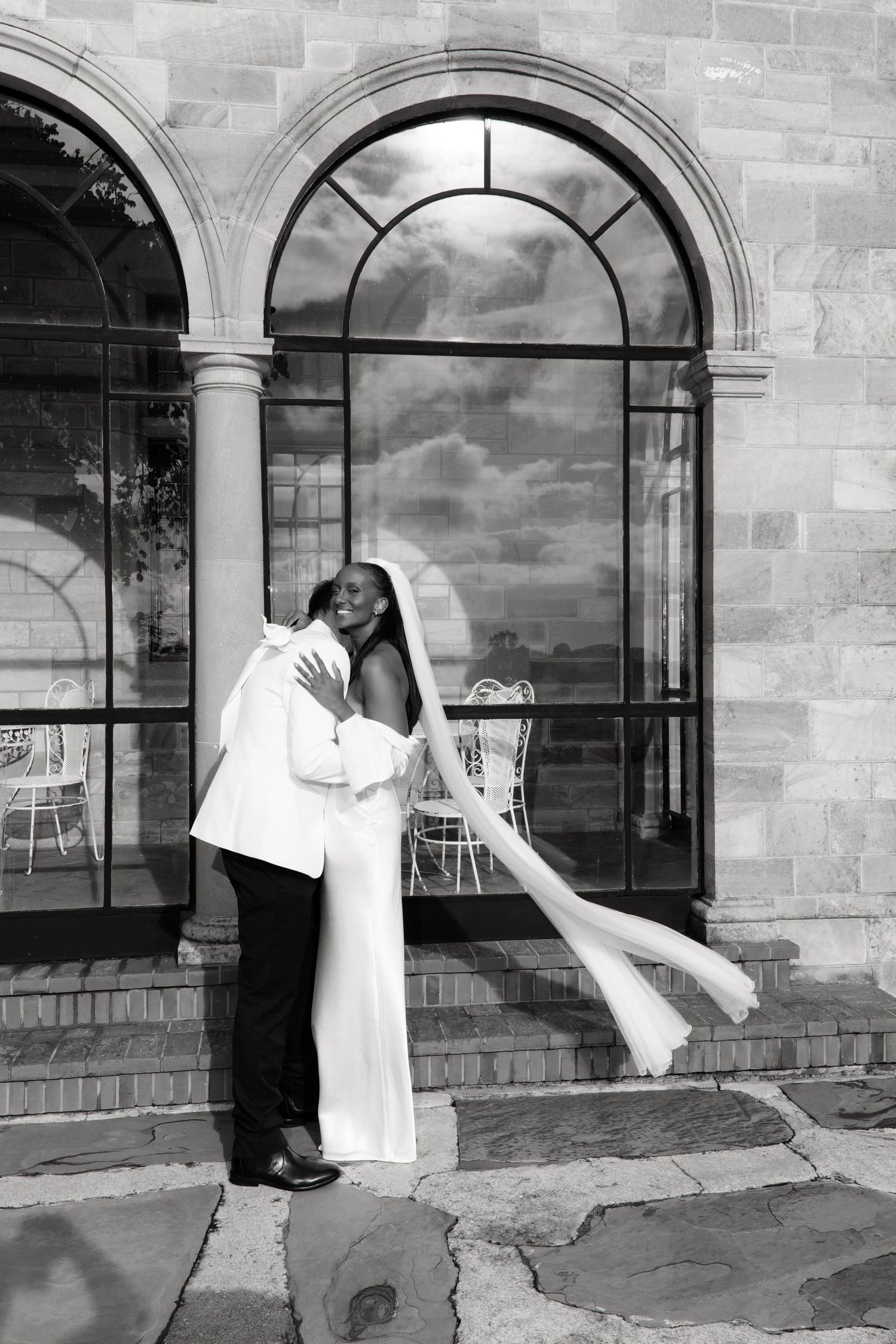 Bride and groom share a romantic embrace outside a stone mansion in black and white.
