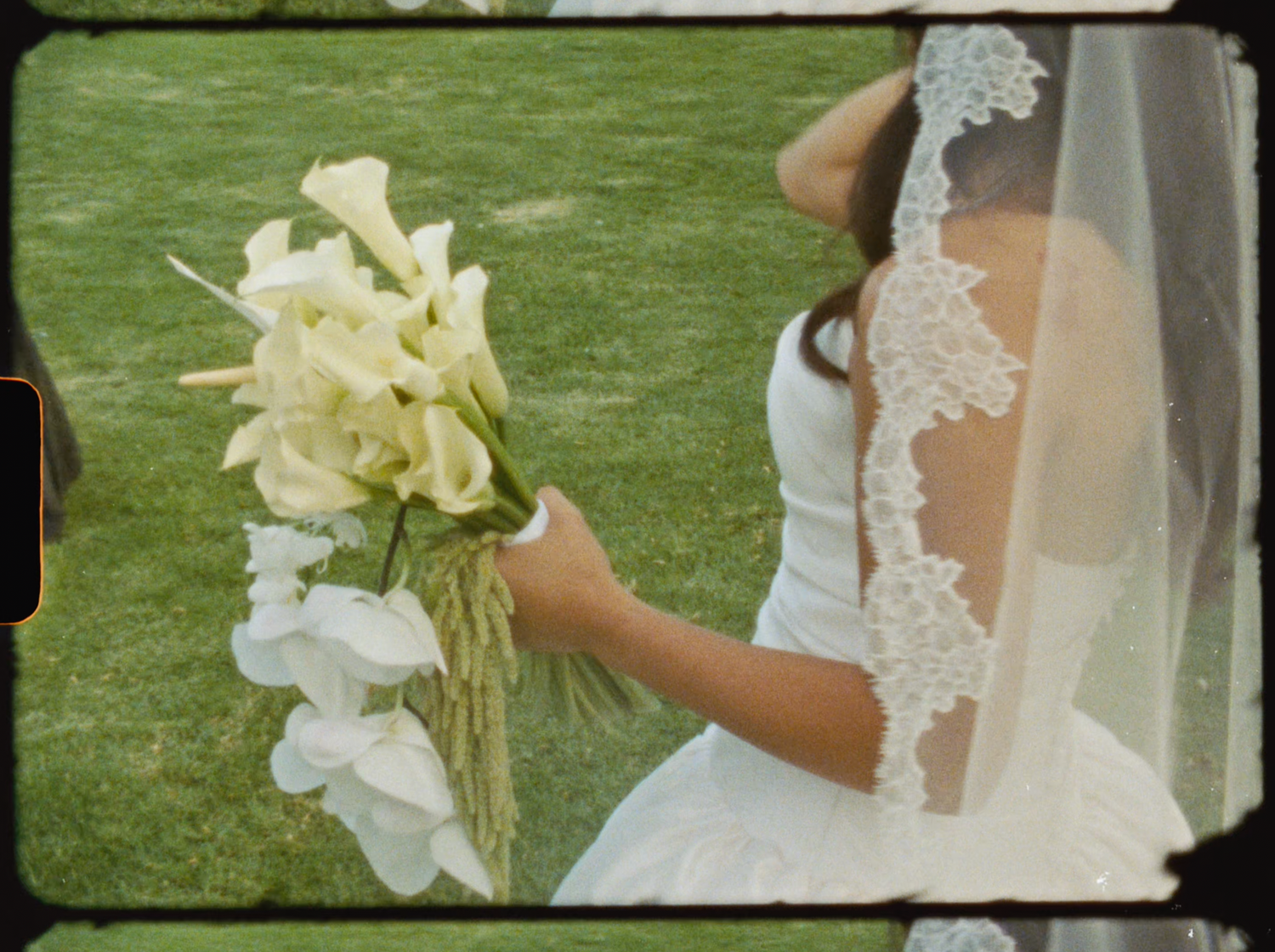 A bride in a white dress and lace veil holds a bouquet of white calla lilies on a green lawn.