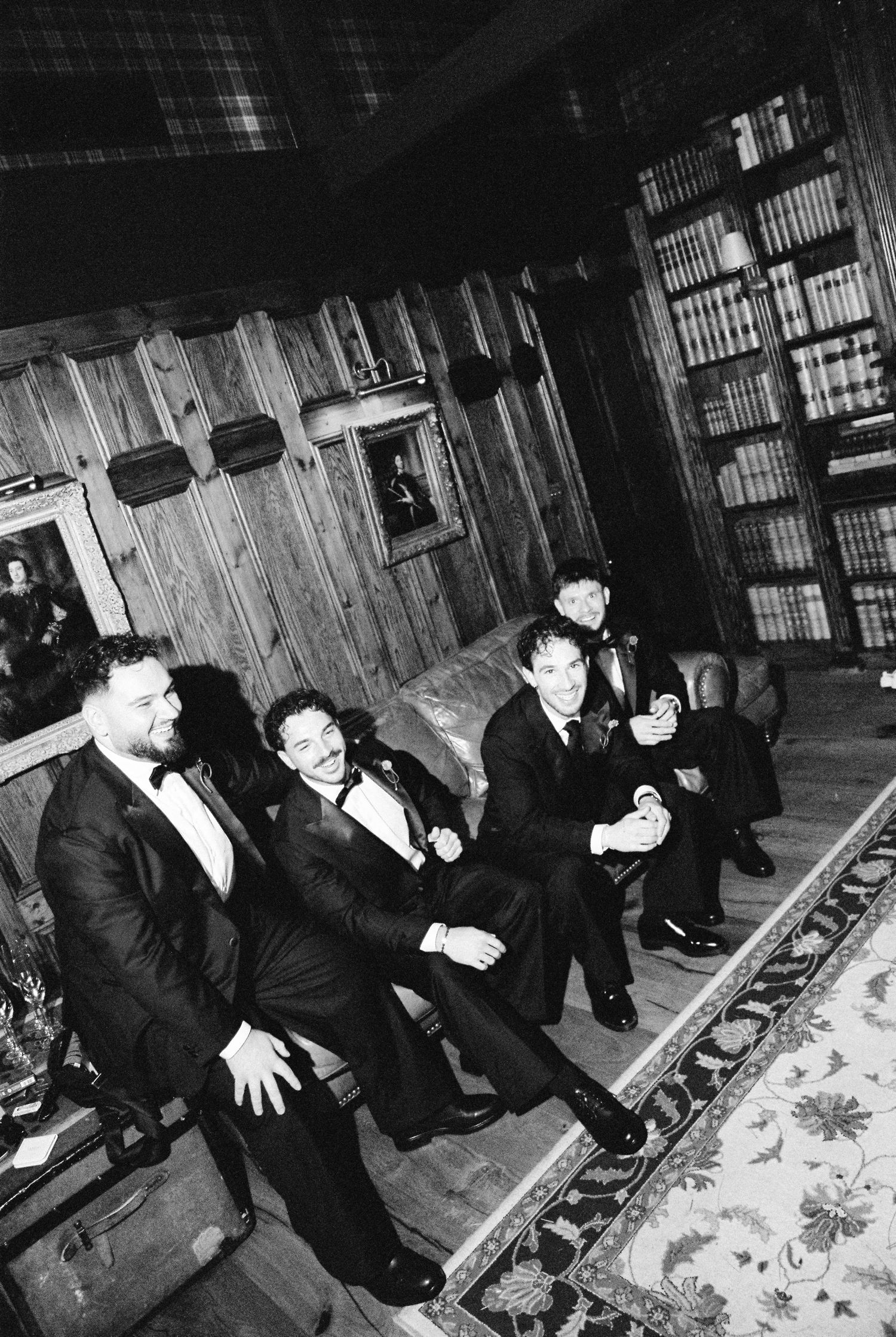 Black and white photo of groomsmen in tuxedos laughing and relaxing in a wood-paneled library at a wedding reception.