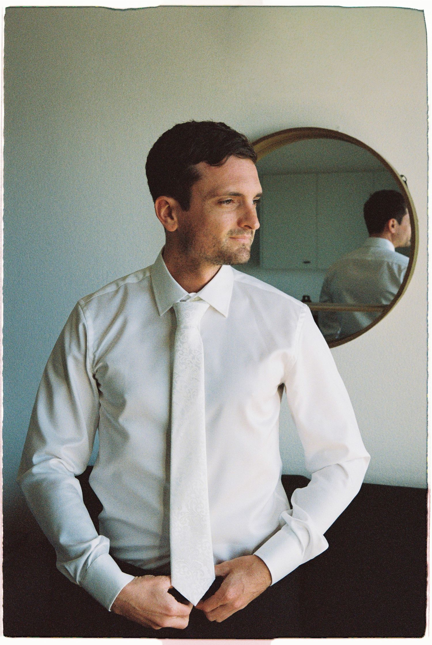 Groom in white dress shirt and tie adjusting outfit before wedding with round mirror behind him.