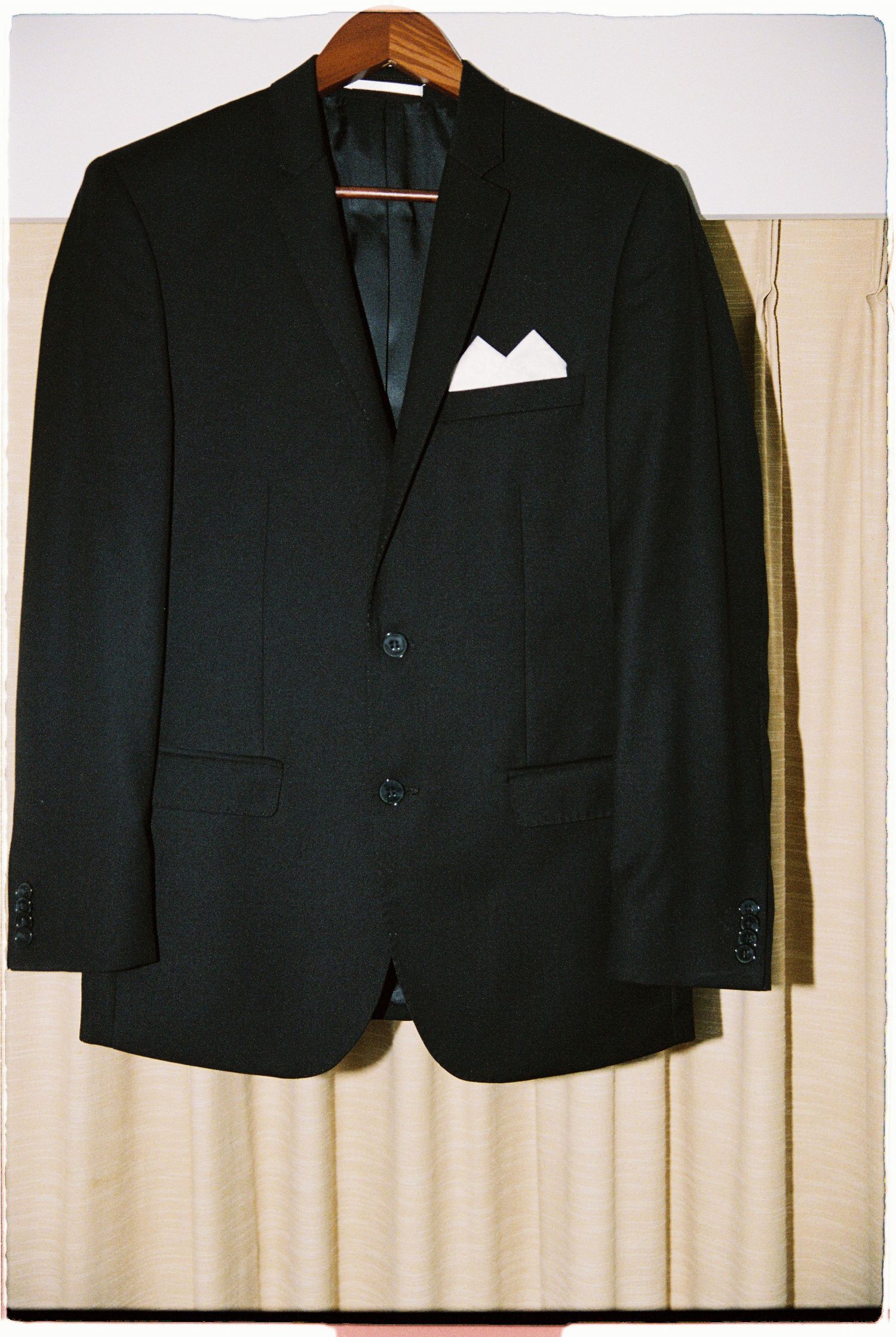 Black two-button suit jacket with white pocket square on wooden hanger against beige curtain.