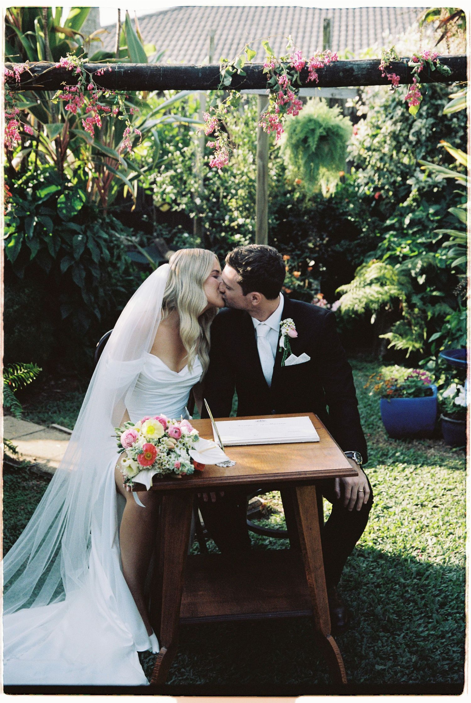 Newlyweds share a kiss at signing table with pink bouquet under floral arch in lush garden setting.