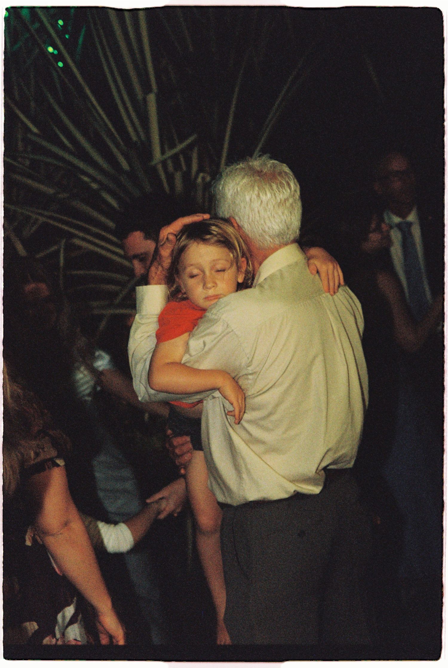 An elderly man tenderly holds a sleeping child at an outdoor evening wedding reception.