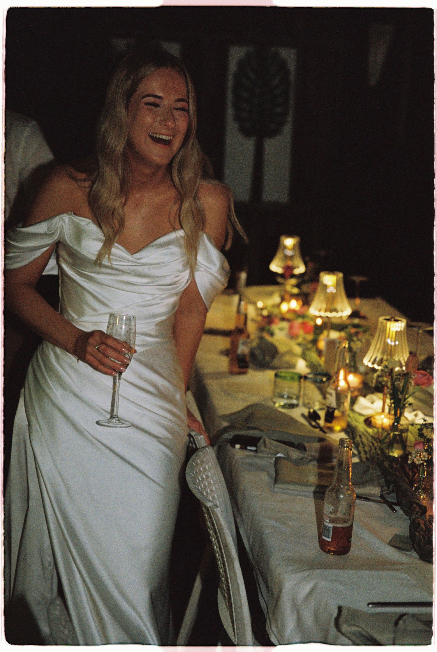 A laughing bride in a white off-shoulder satin gown holds a champagne flute at her candlelit wedding reception.