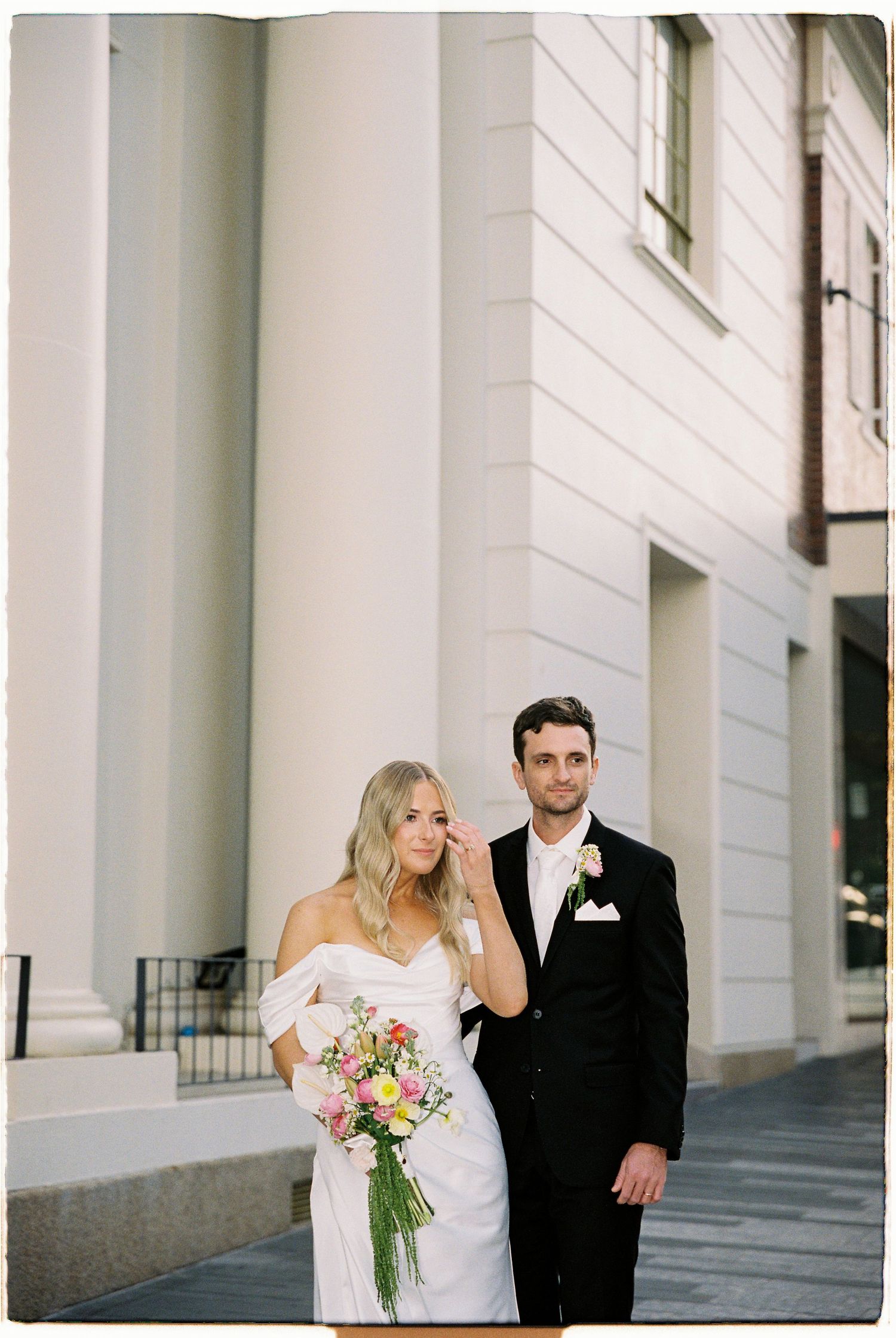 Smiling bride holding colorful bouquet stands with groom beside white neoclassical building columns.