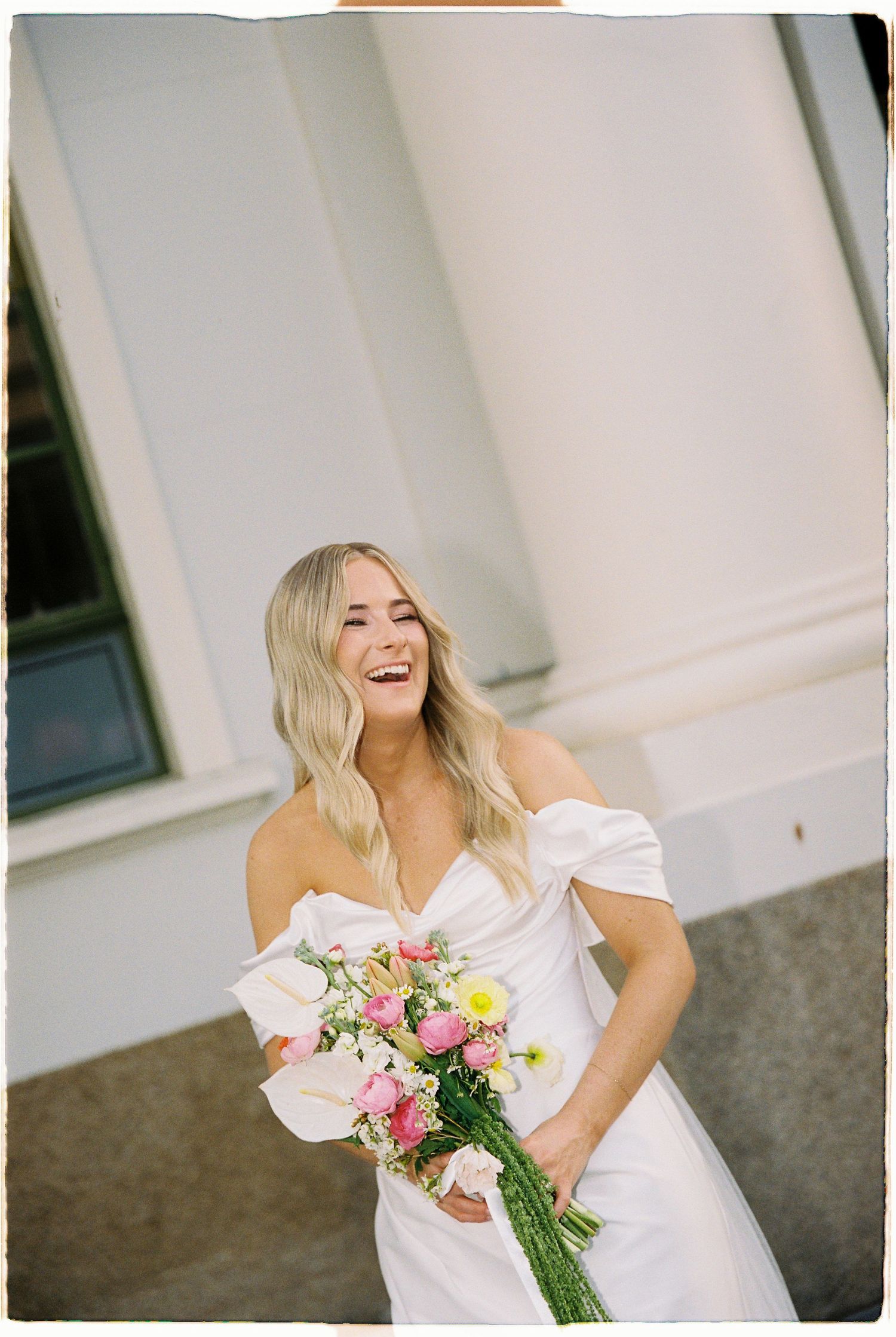 Joyful blonde bride laughs while holding vibrant pink and yellow floral bouquet in white gown.