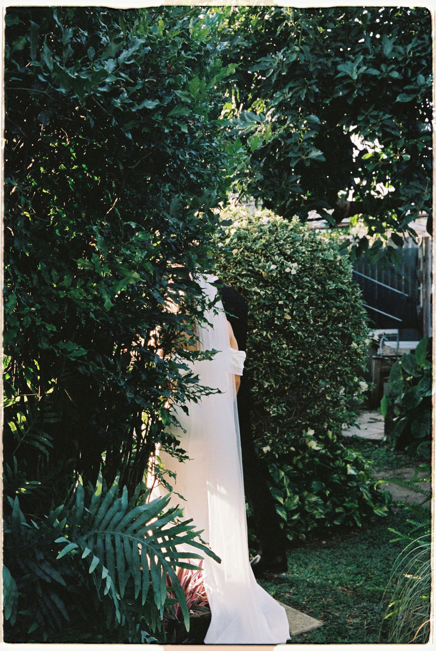 Bride in flowing white wedding gown and cathedral veil stands among lush tropical garden greenery.