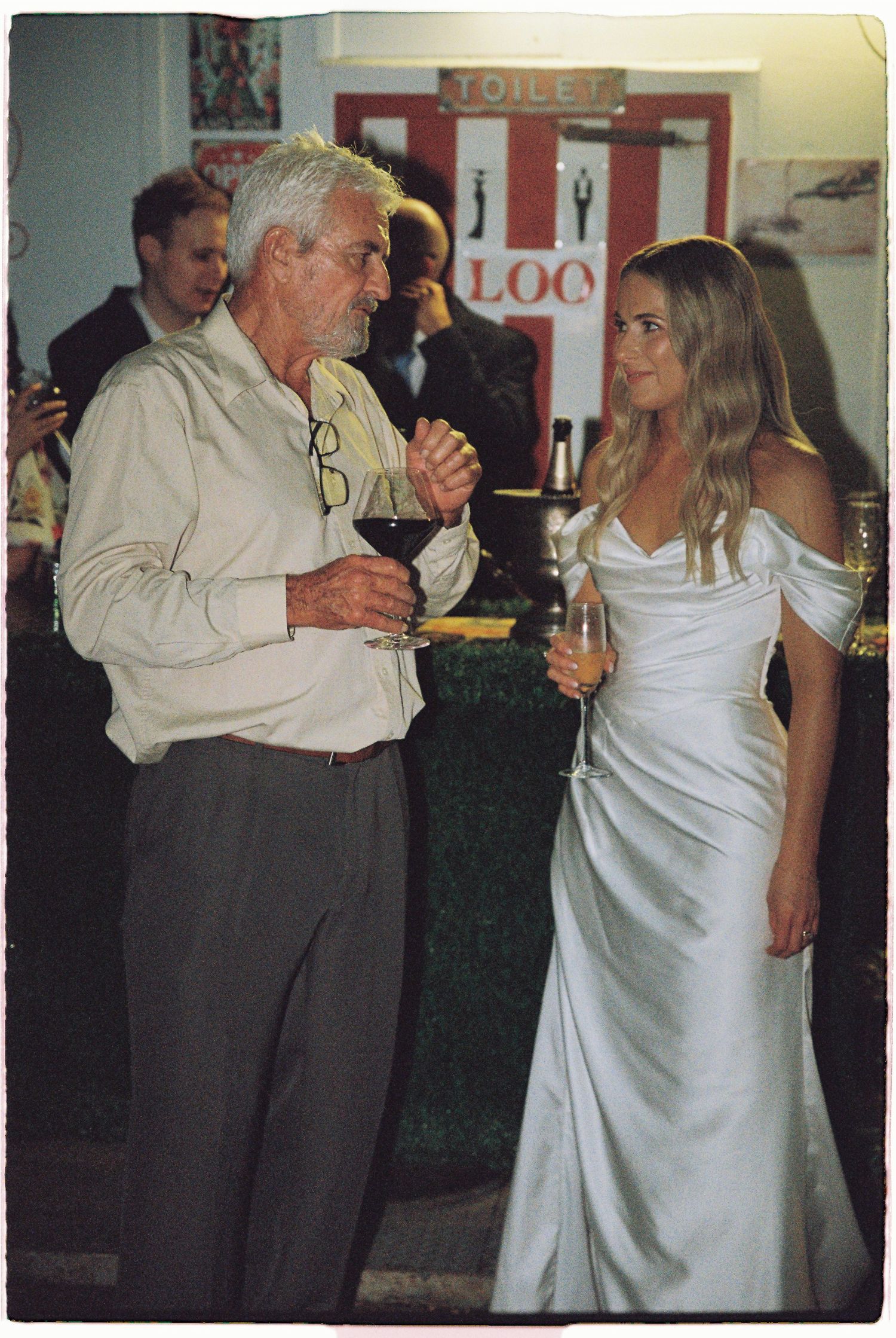 Bride in off-shoulder white gown converses with older gentleman holding wine glass at indoor reception.