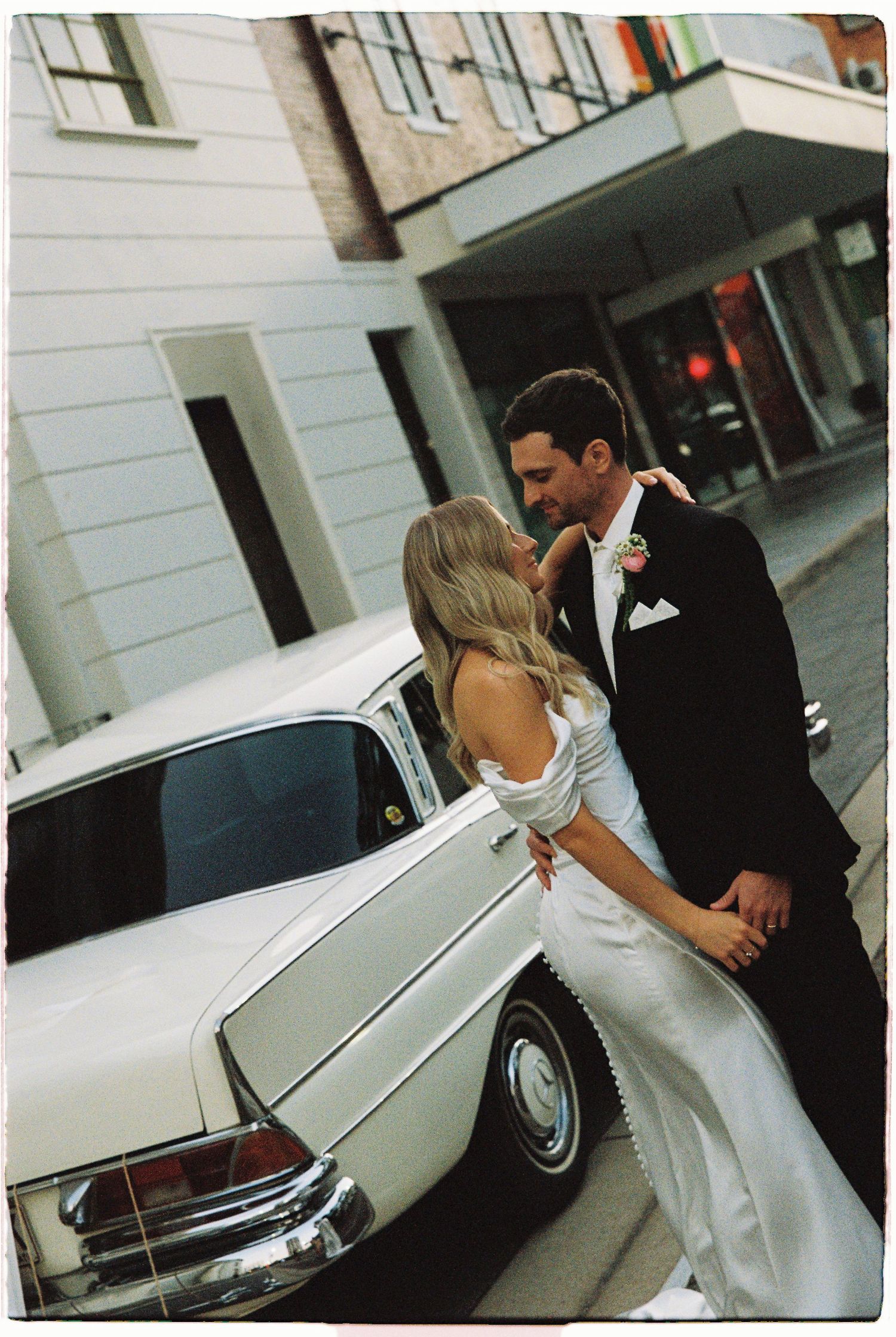 Bride and groom share romantic kiss leaning against a classic white vintage car on city street.