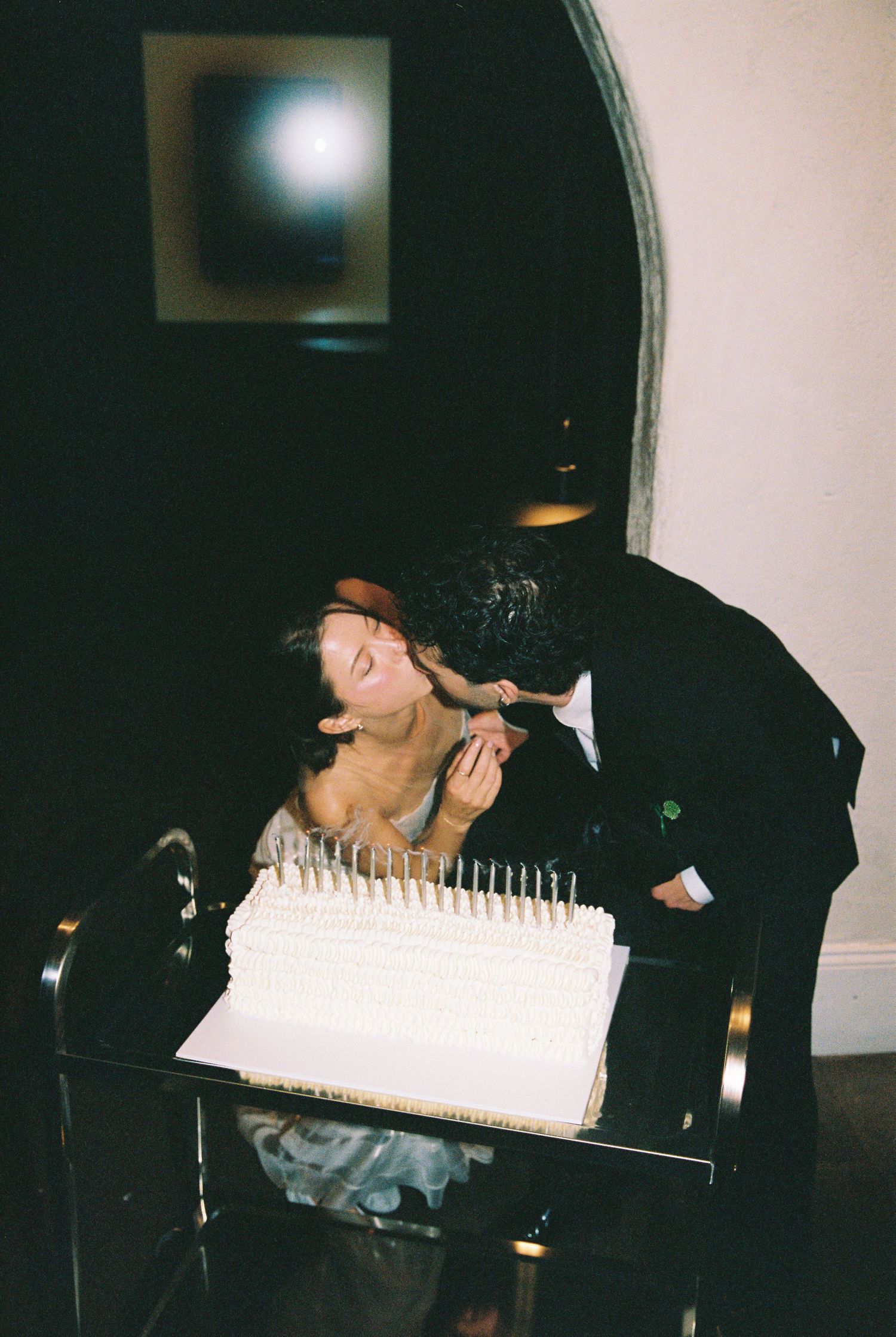 Bride and groom sharing a romantic kiss over their white wedding cake adorned with tall candles.