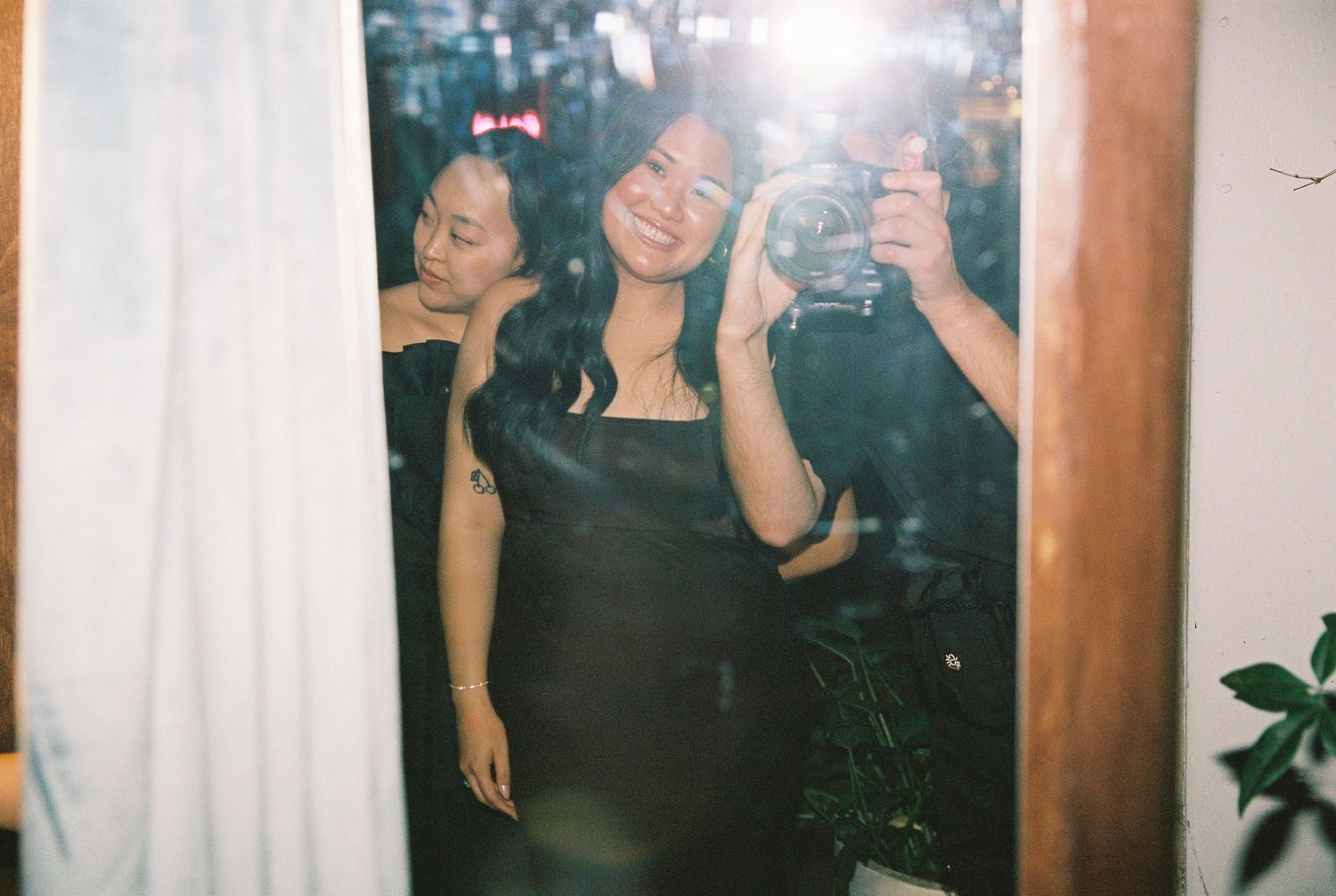 Two women posing for a mirror selfie at a party, one in a black dress smiling at the camera.