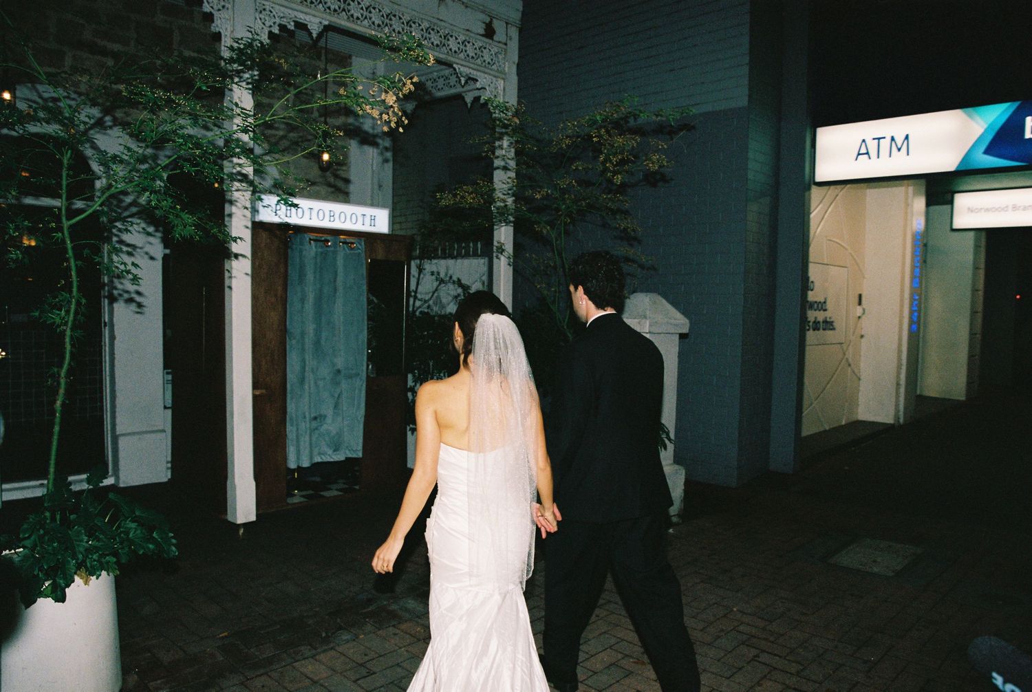 Bride in white mermaid gown and groom walking hand in hand past an ATM at night.