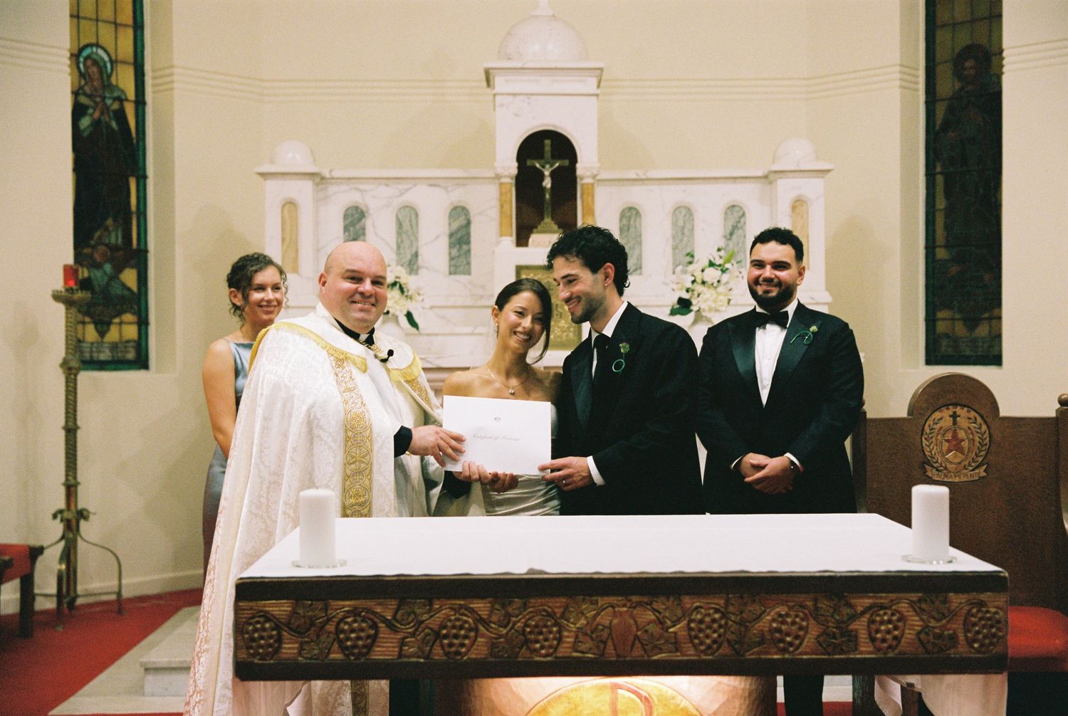 Priest presenting wedding certificate to smiling bride and groom at church altar ceremony.