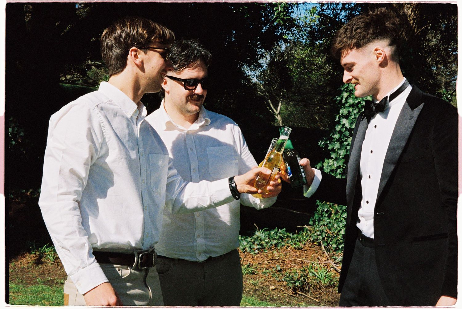 Three men in formal attire toasting with beer bottles outdoors in a garden setting.