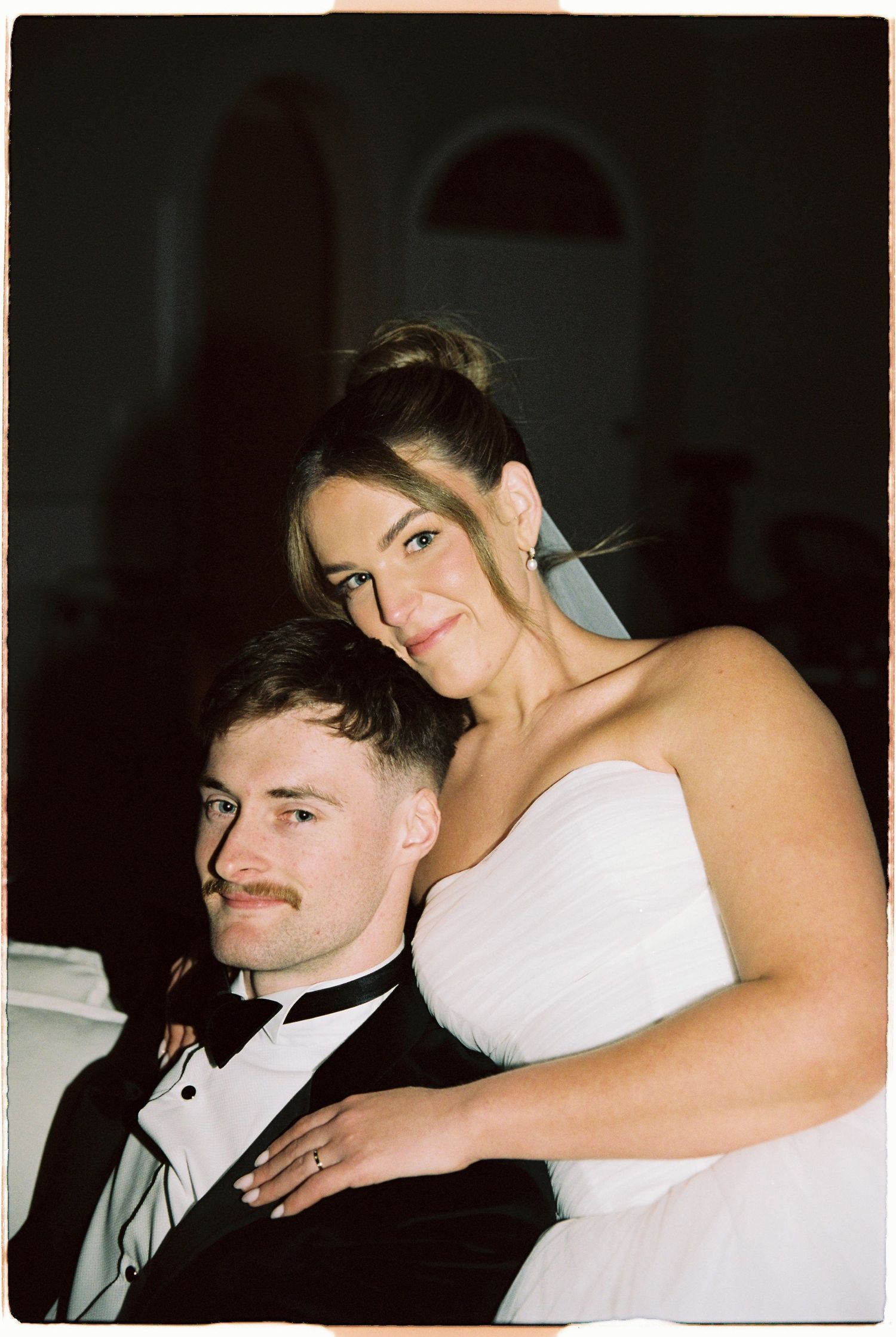 Bride and groom posing together at wedding reception, groom in black tuxedo.