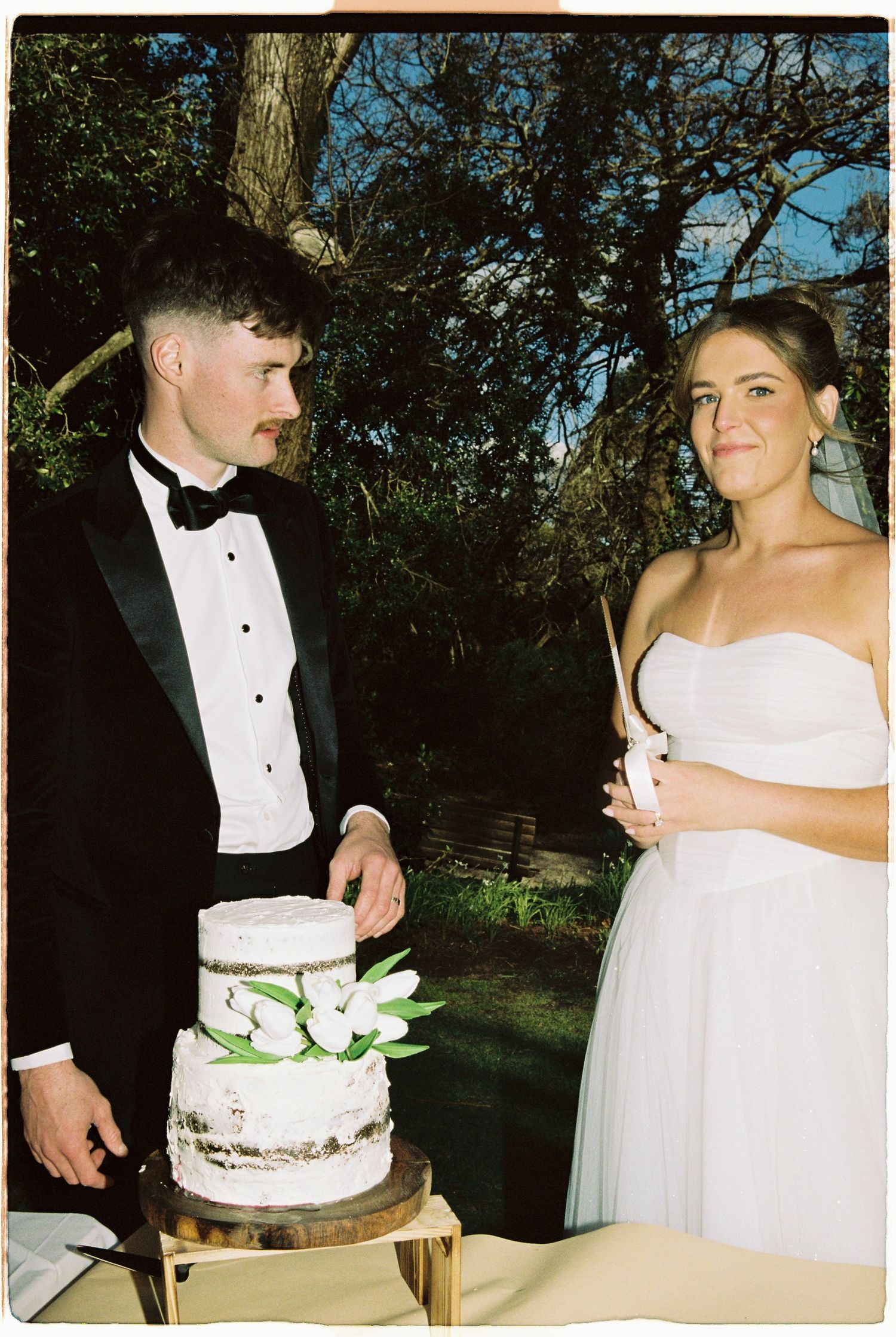 Bride and groom posing beside a rustic naked wedding cake decorated with white tulips.