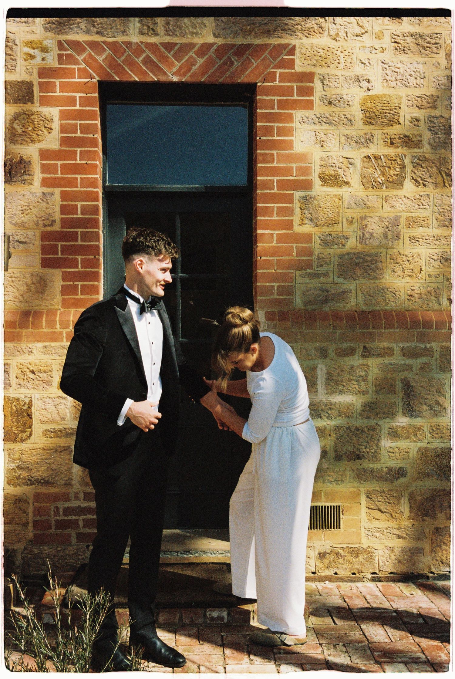 Groom in tuxedo sharing a tender moment with bride in white dress outside stone building.