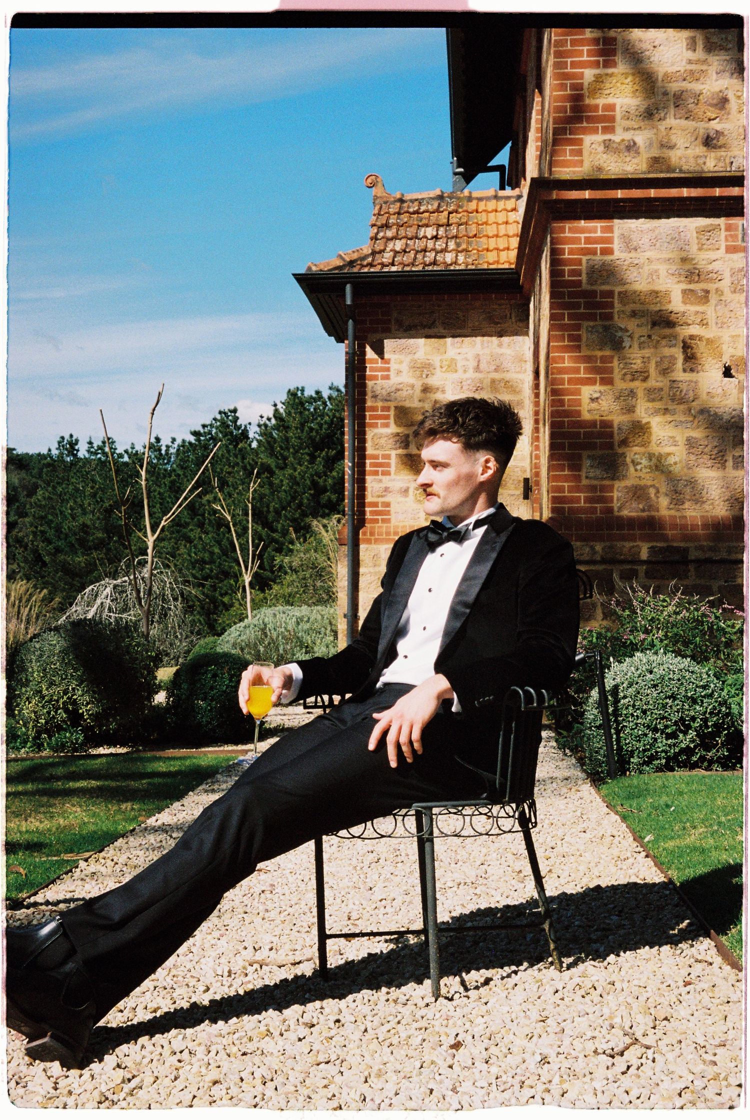 A young man in a tuxedo relaxes on a garden chair holding a drink outside a brick building on a sunny day.