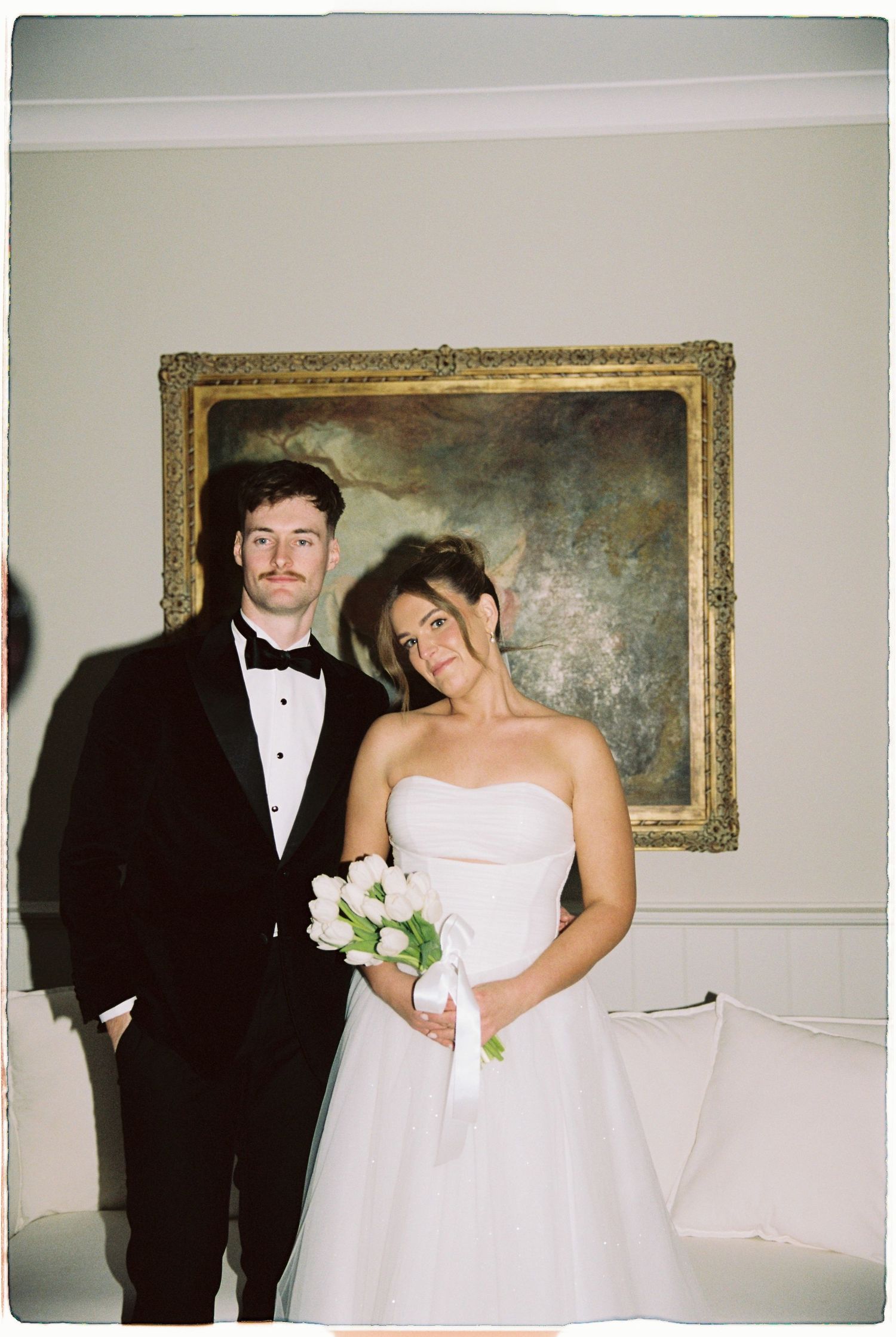 Bride and groom posing indoors before a gold-framed painting, groom in tuxedo, bride holding white bouquet.
