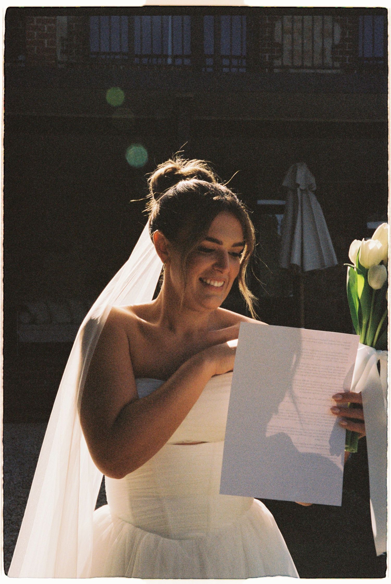 Smiling bride in white strapless gown and veil reading vows in sunlit outdoor ceremony.