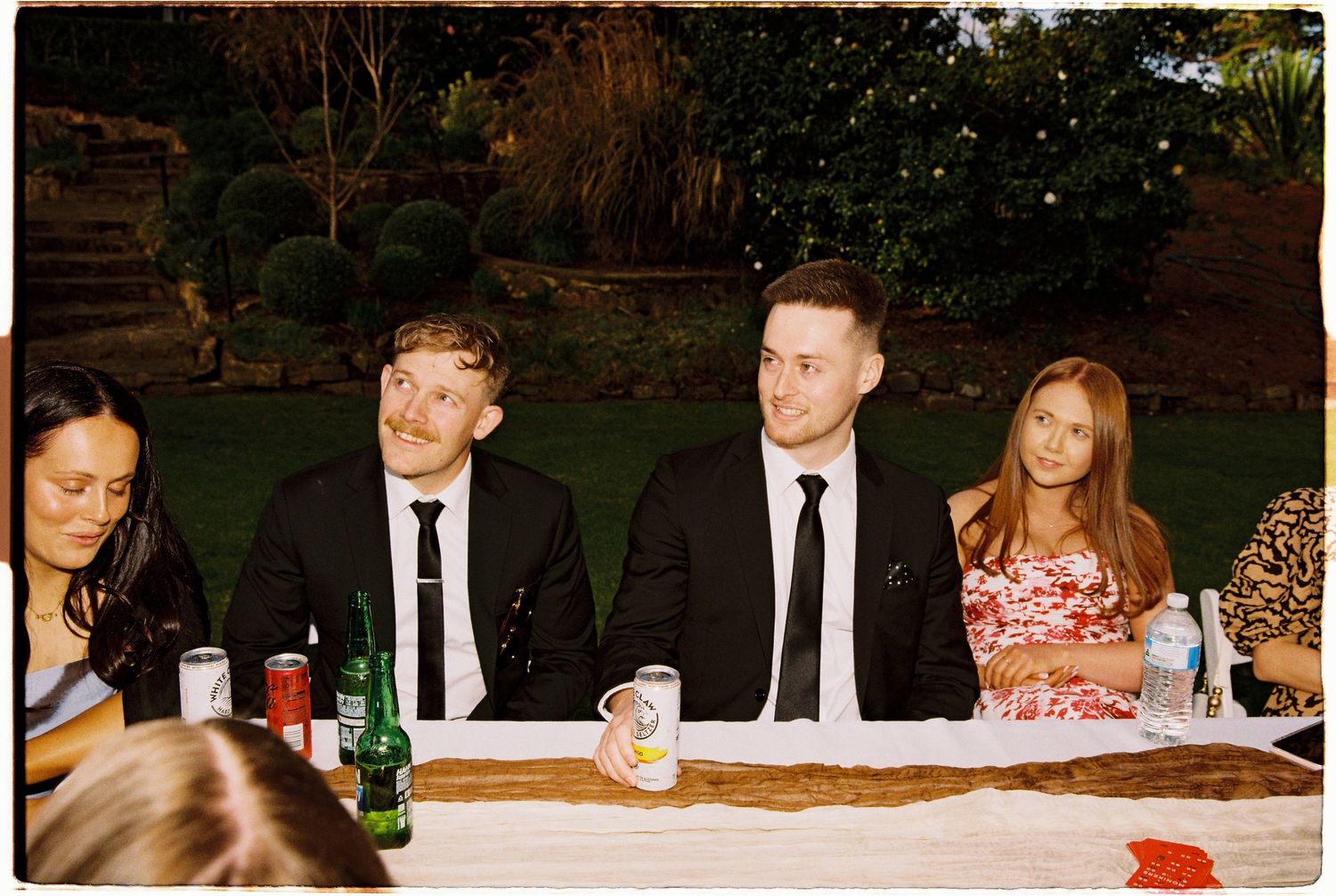 Wedding guests in formal attire seated at an outdoor reception dinner table with drinks.