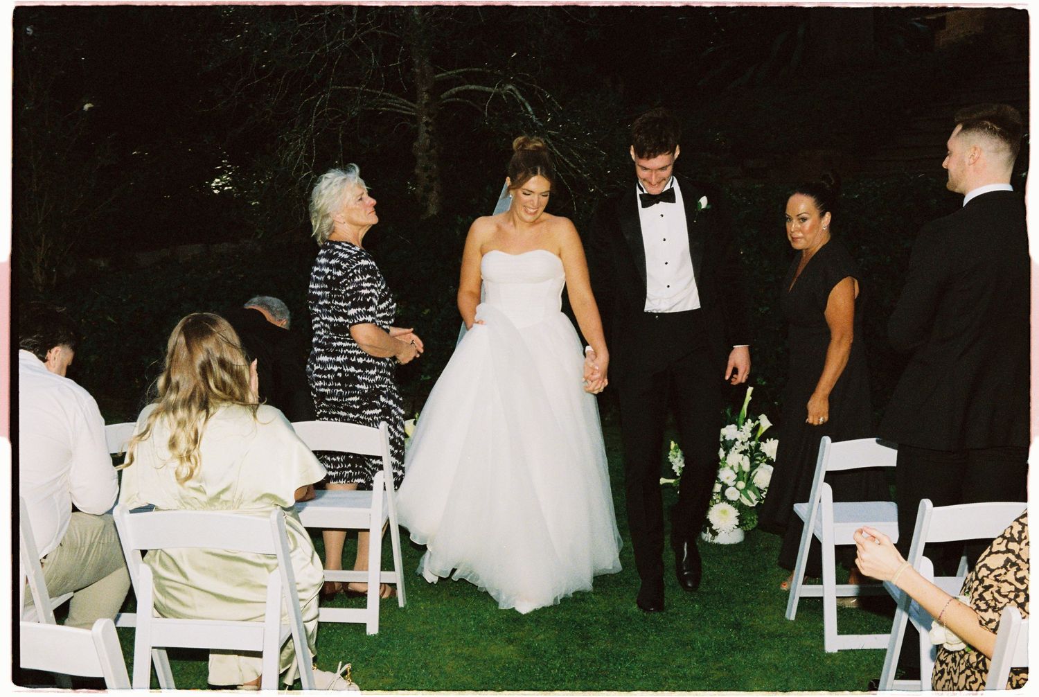 Bride and groom walk down the aisle at an outdoor evening wedding ceremony with guests seated on white chairs.