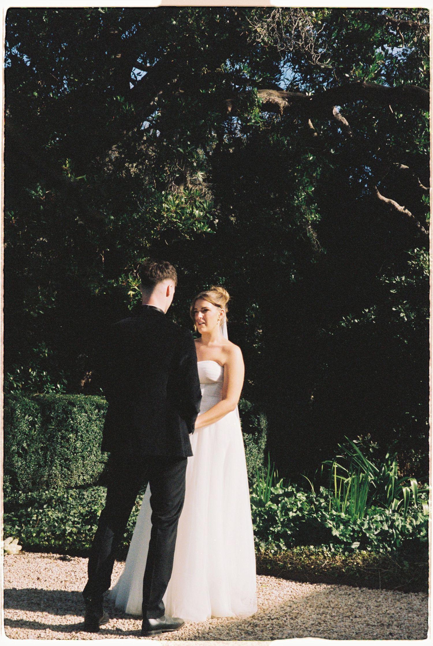 Bride in white strapless gown and groom in black suit sharing a moment in a lush garden setting.