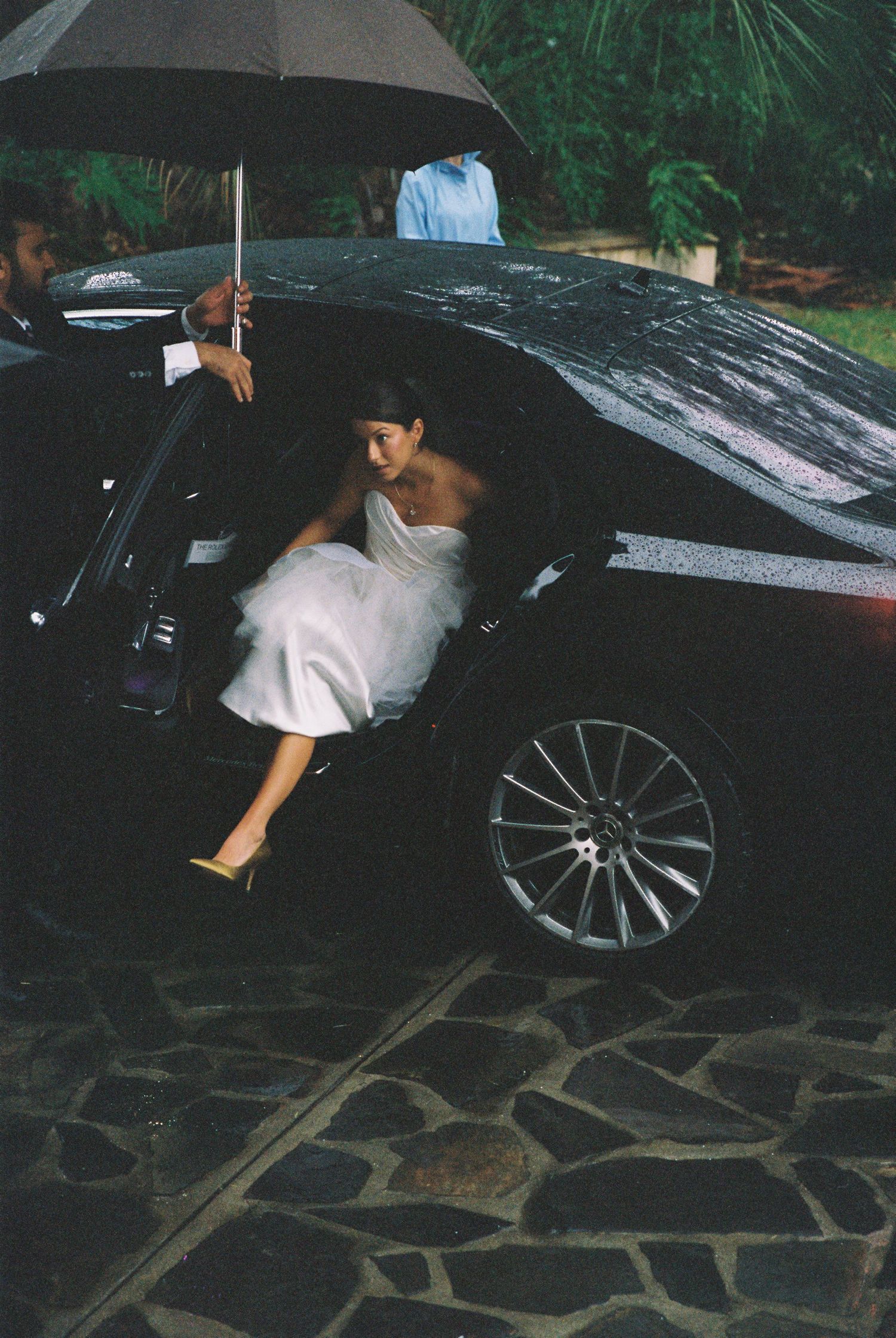 Bride in white strapless gown stepping out of black car under umbrella on rainy wedding day.