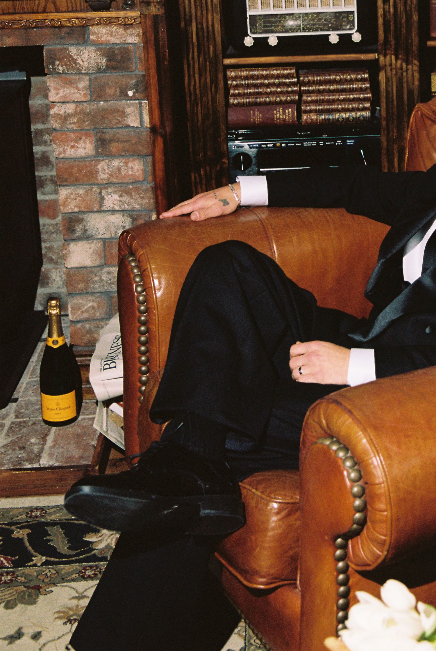 Groom in black tuxedo relaxing in leather armchair beside Veuve Clicquot champagne bottle.