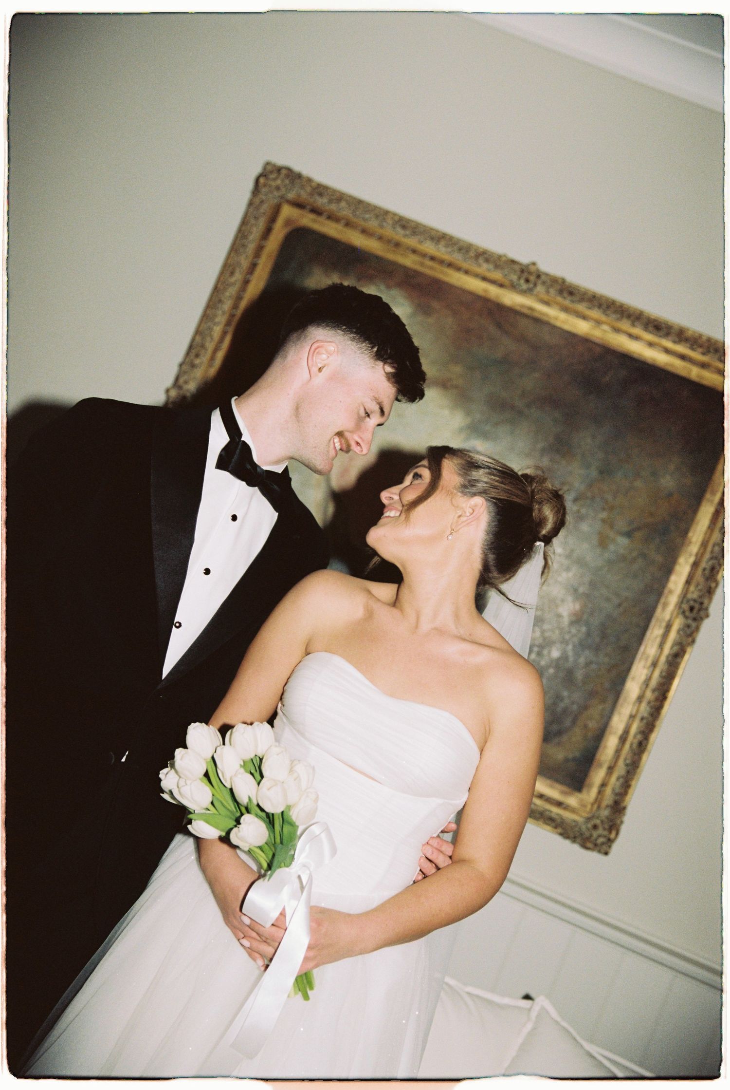 Bride holding white tulip bouquet and groom in tuxedo share a joyful moment before a classic painting.