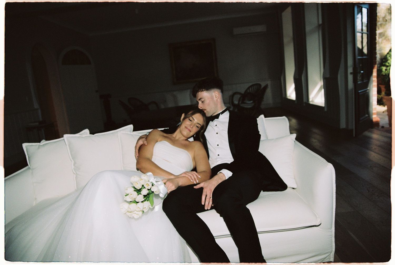 Bride and groom in wedding attire relax together on white sofa in elegant interior setting.
