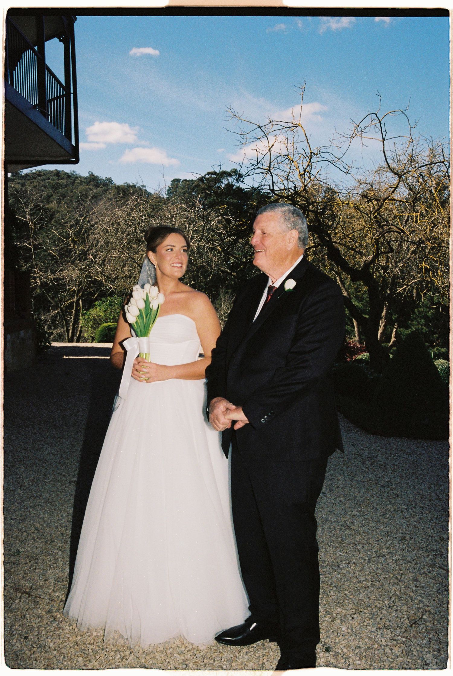 Bride in white strapless gown holding tulip bouquet stands with older man in black suit at outdoor wedding venue.