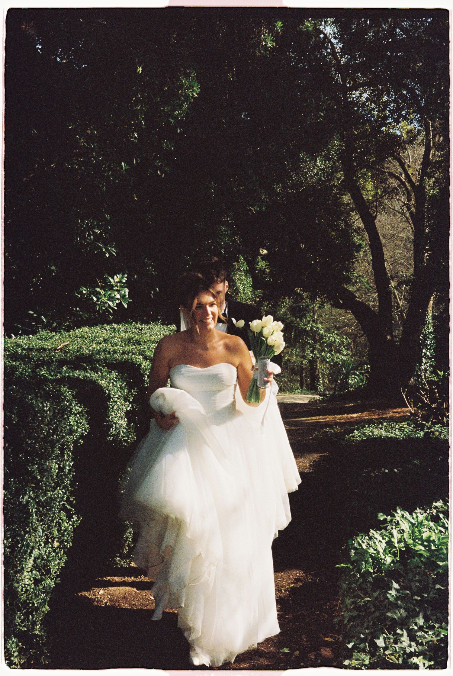 Bride in a strapless white ball gown walks along a garden path holding a white bouquet of flowers.