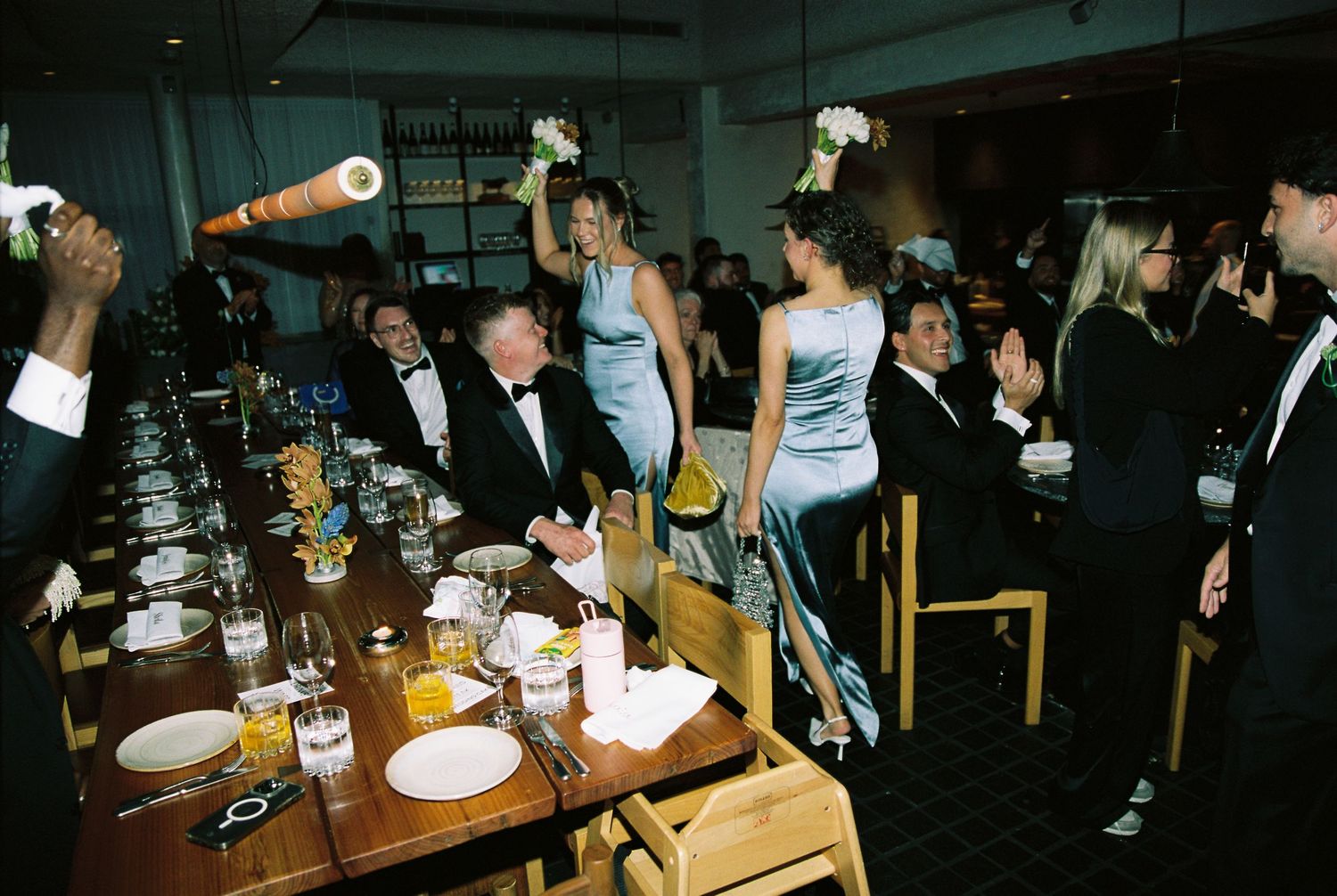 Wedding guests cheer and celebrate along a long decorated reception dinner table.